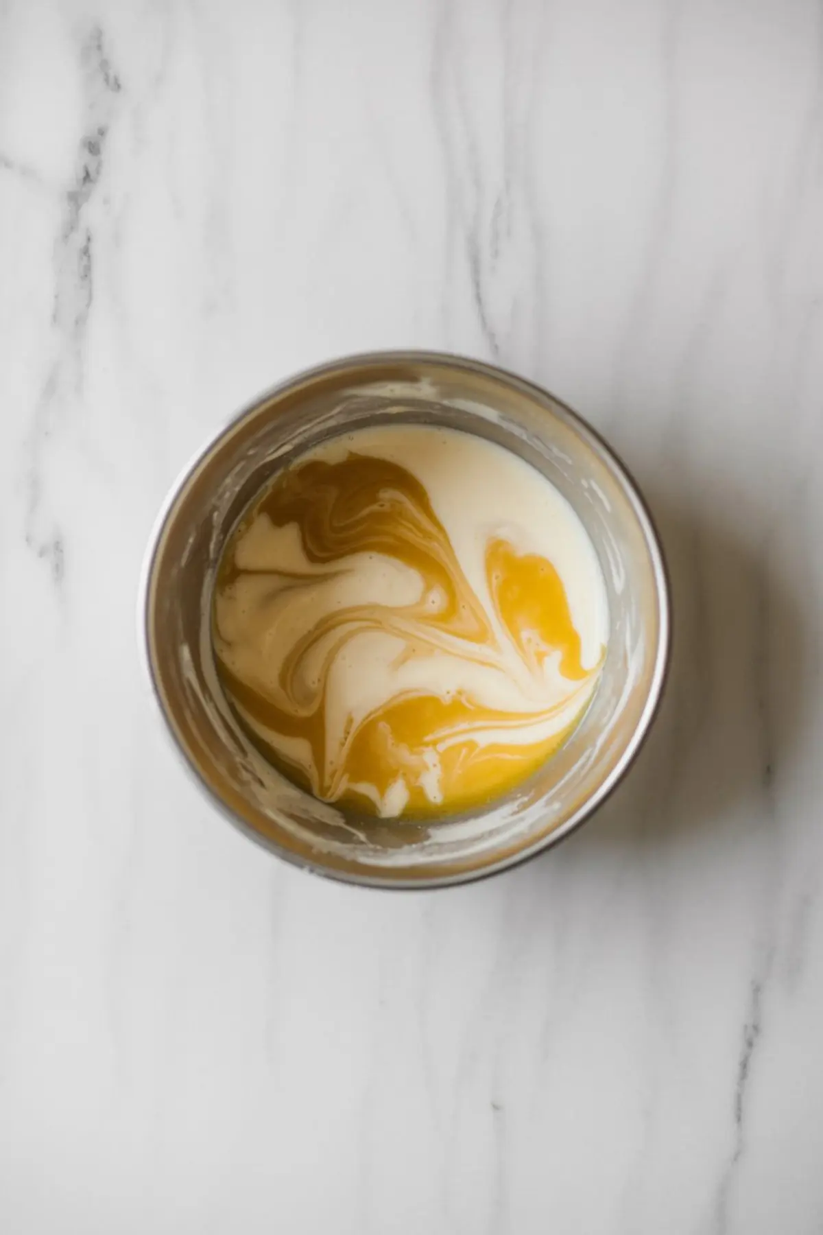 Swirled mixture of melted butter, milk, and vanilla extract in a stainless steel mixing bowl, showing marbled patterns on a white countertop before combining with dry ingredients.