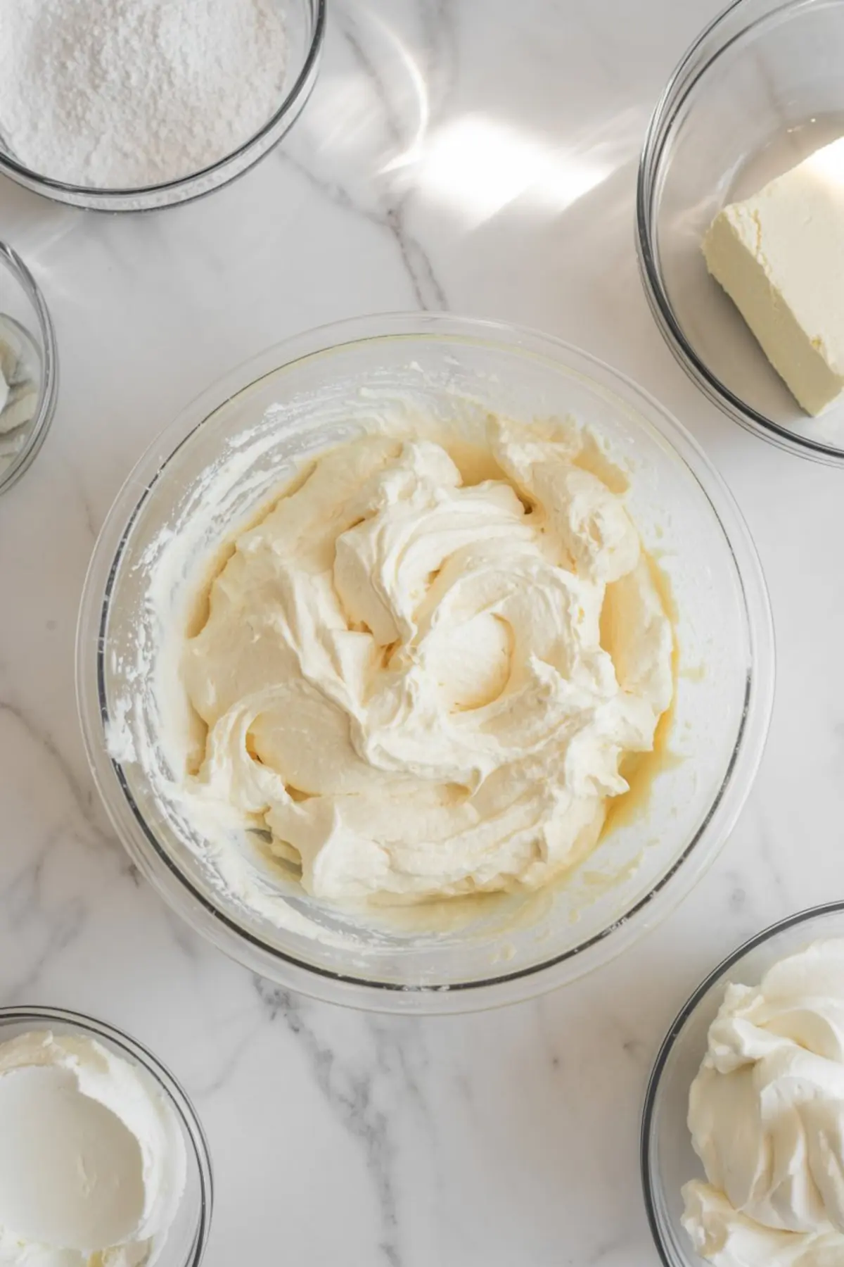 Creamy cheesecake filling whipped in a glass mixing bowl, surrounded by ingredients including powdered sugar, cream cheese, and whipped topping on a white marble counter.
