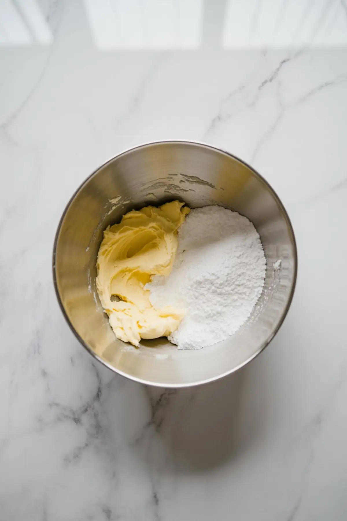 Butter and powdered sugar in a stainless steel mixing bowl, placed on a white marble countertop, ready for making cookie dough.
