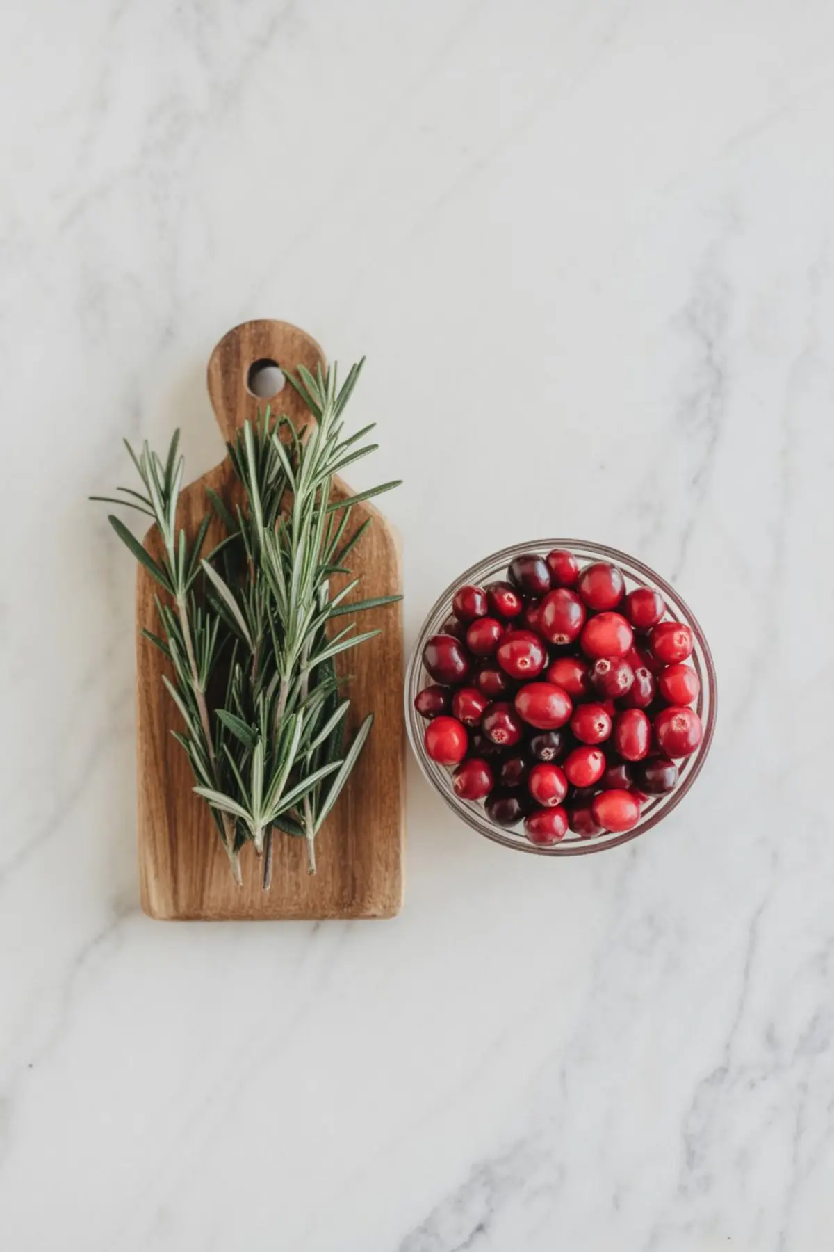 Flat lay of fresh rosemary on a wooden cutting board next to a glass bowl filled with whole cranberries on white marble. These natural ingredients are used for making festive holiday ice cubes.

