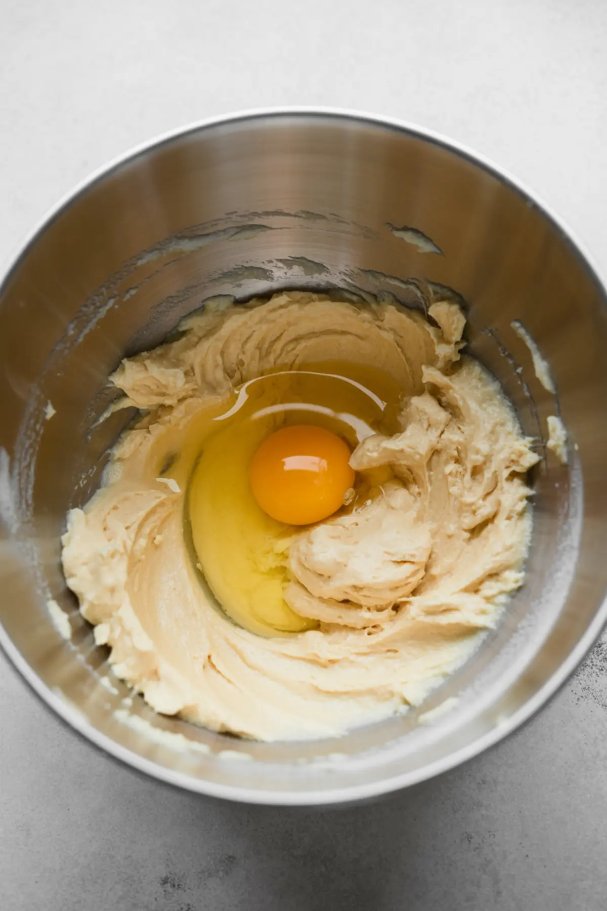 A metal mixing bowl with creamy butter mixture and a raw egg added in the center during the cake batter mixing process.
