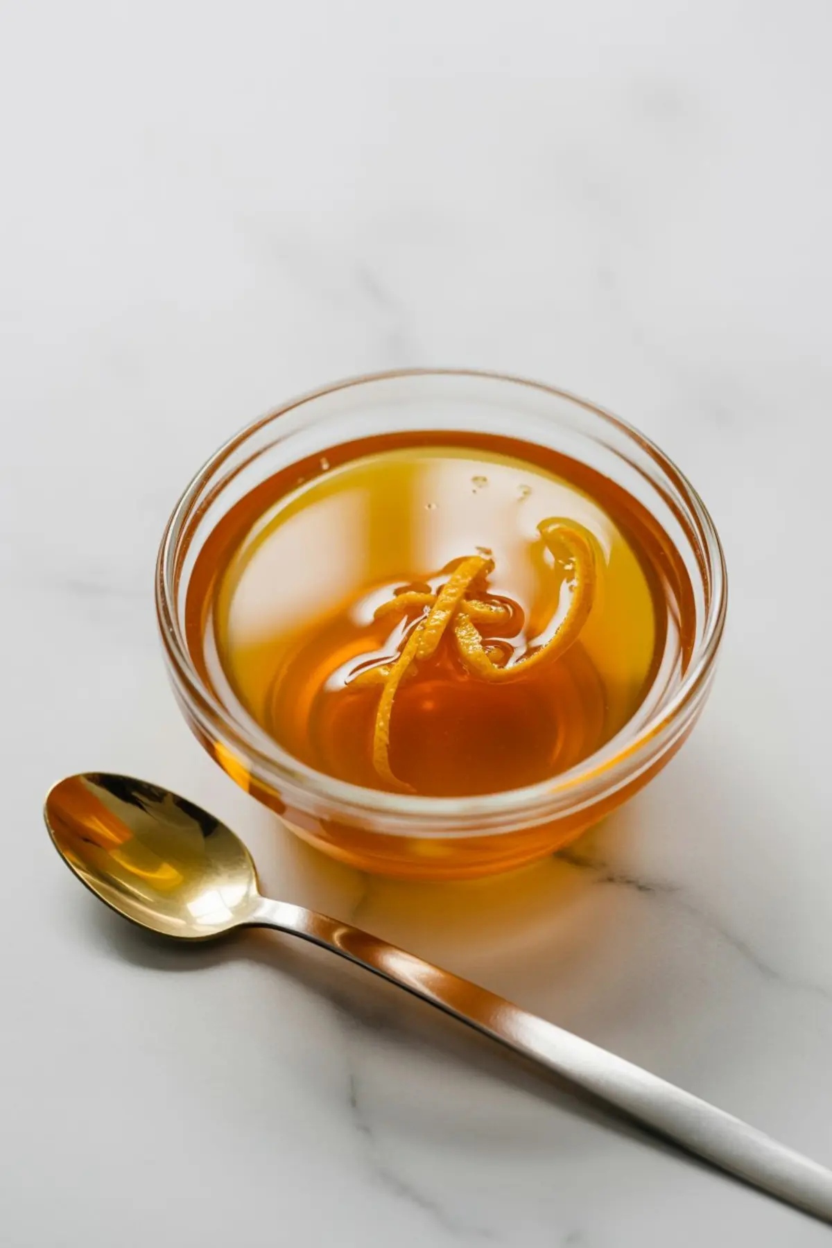 A small glass bowl filled with amber orange syrup and thin orange zest strips beside a gold spoon on a marble surface.
