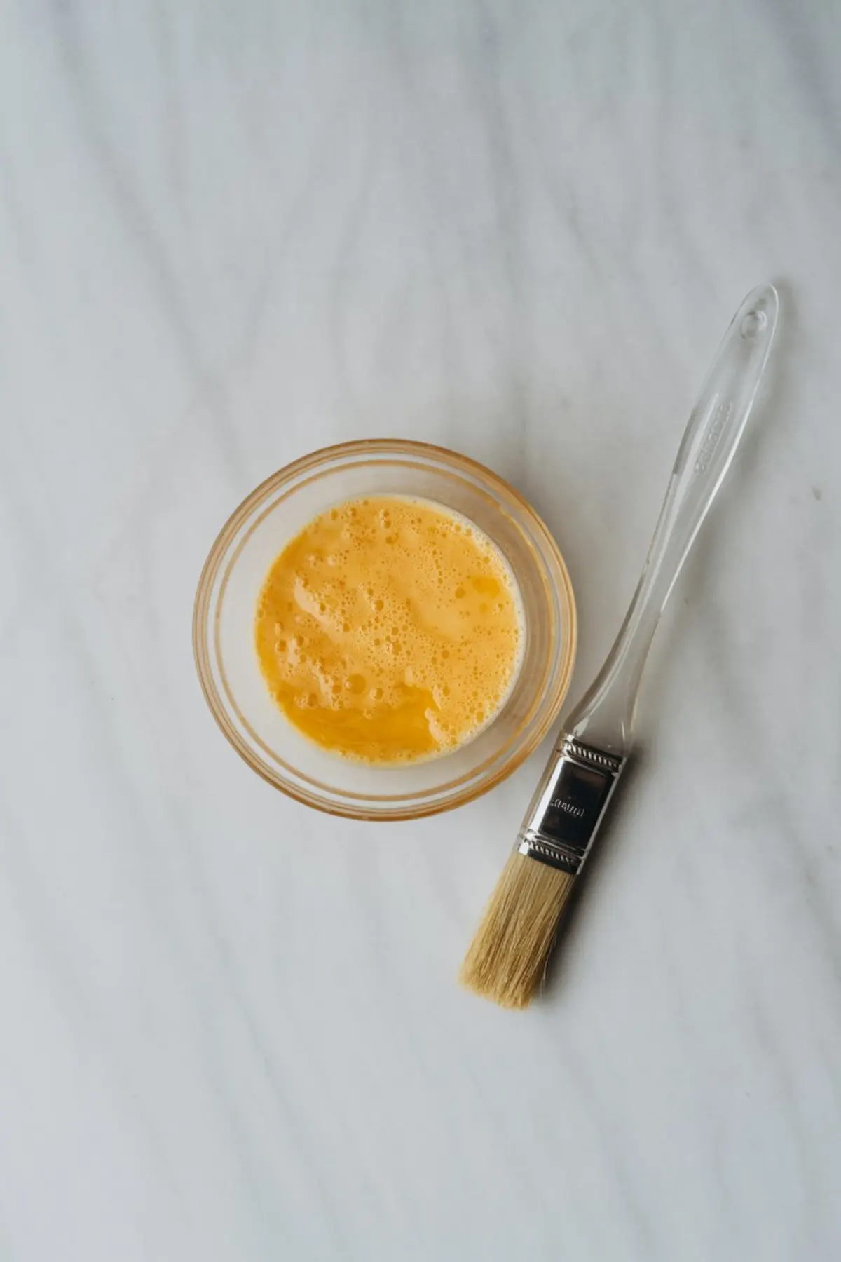 Small glass bowl filled with beaten egg yolk and a pastry brush placed beside it. The mixture is ready for brushing onto pastry dough to give a golden finish during baking, set on a clean marble surface.
