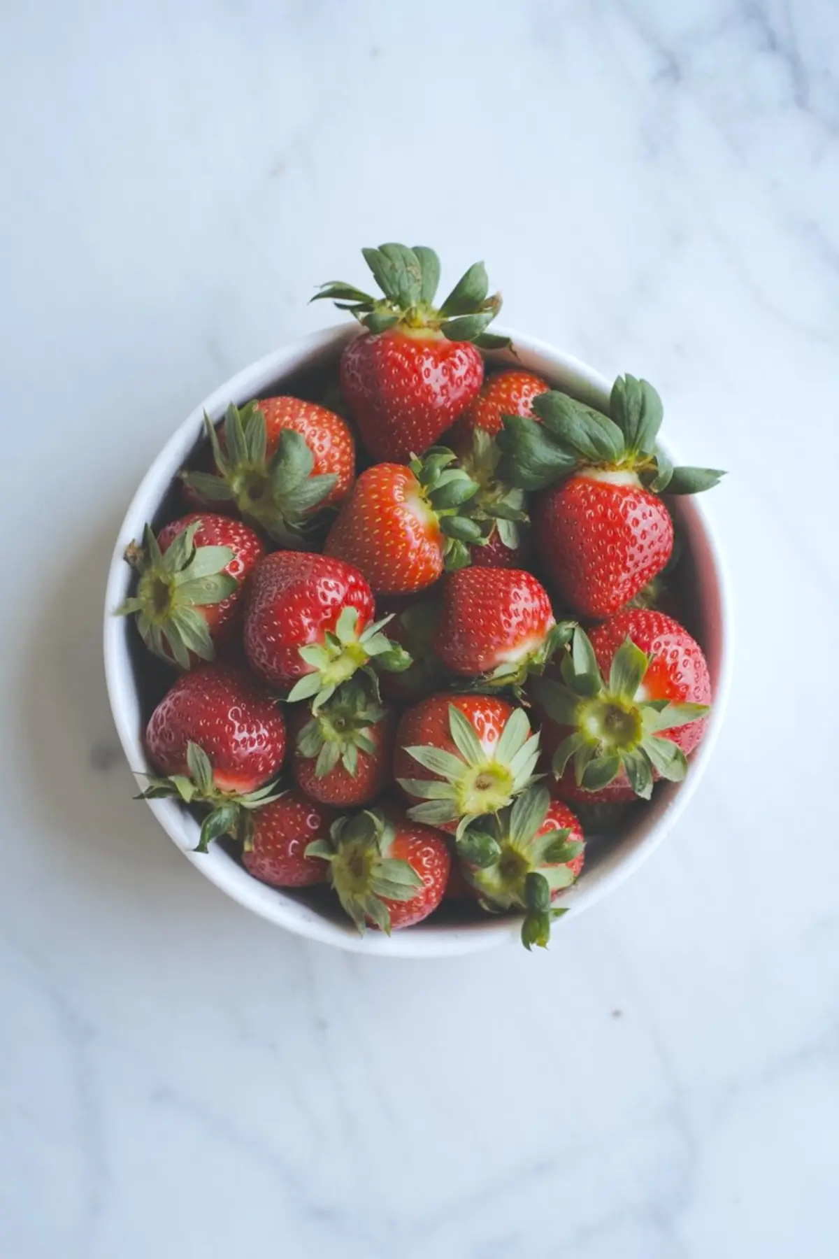 A white bowl filled with vibrant, ripe strawberries with fresh green tops sits on a light marble surface, ideal for dessert recipes or fruit decor ideas.
