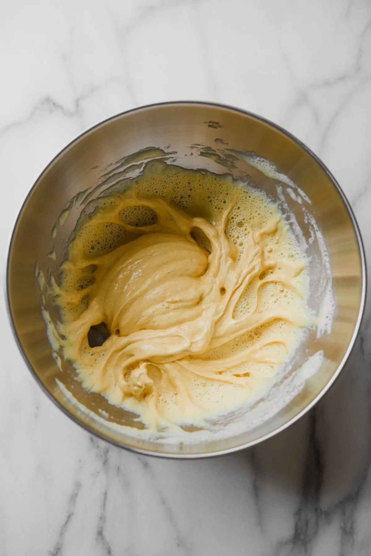Thick whipped batter swirled in a stainless steel mixing bowl on a marble countertop, showing the early stage of making cinnamon maple pecan roulade.