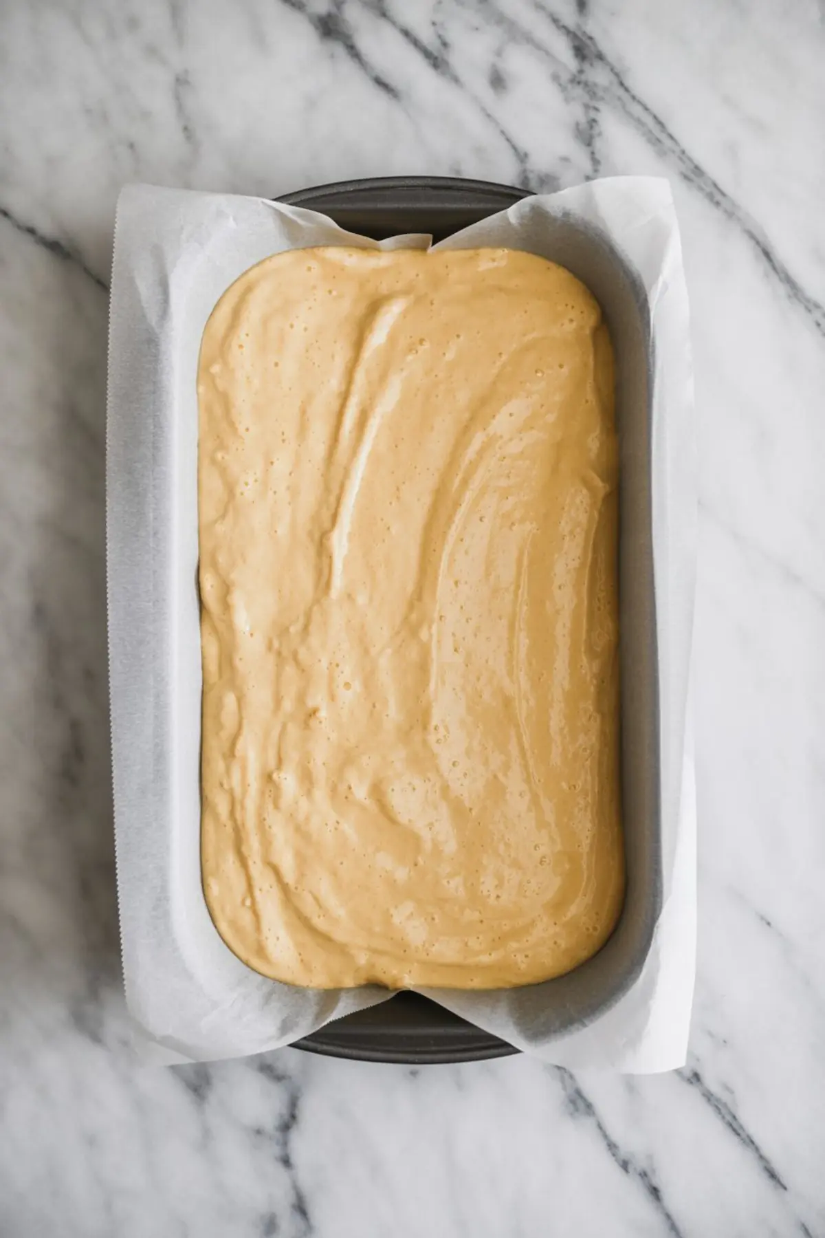 Light golden cake batter spread evenly in a parchment-lined loaf pan, ready for baking on a marble surface.