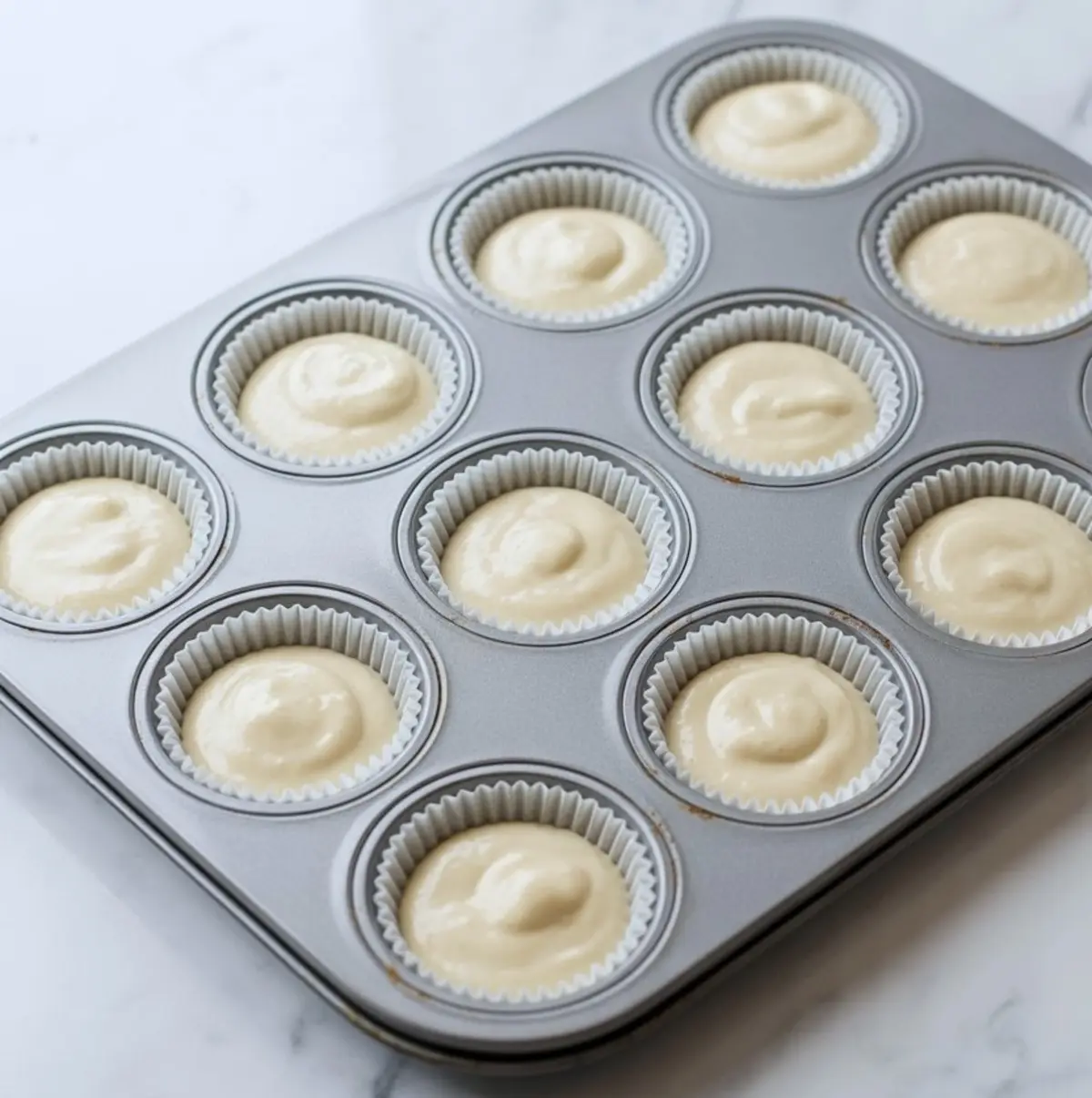 A muffin tin is lined with white cupcake wrappers, each filled with an even portion of cupcake batter, prepared for baking.