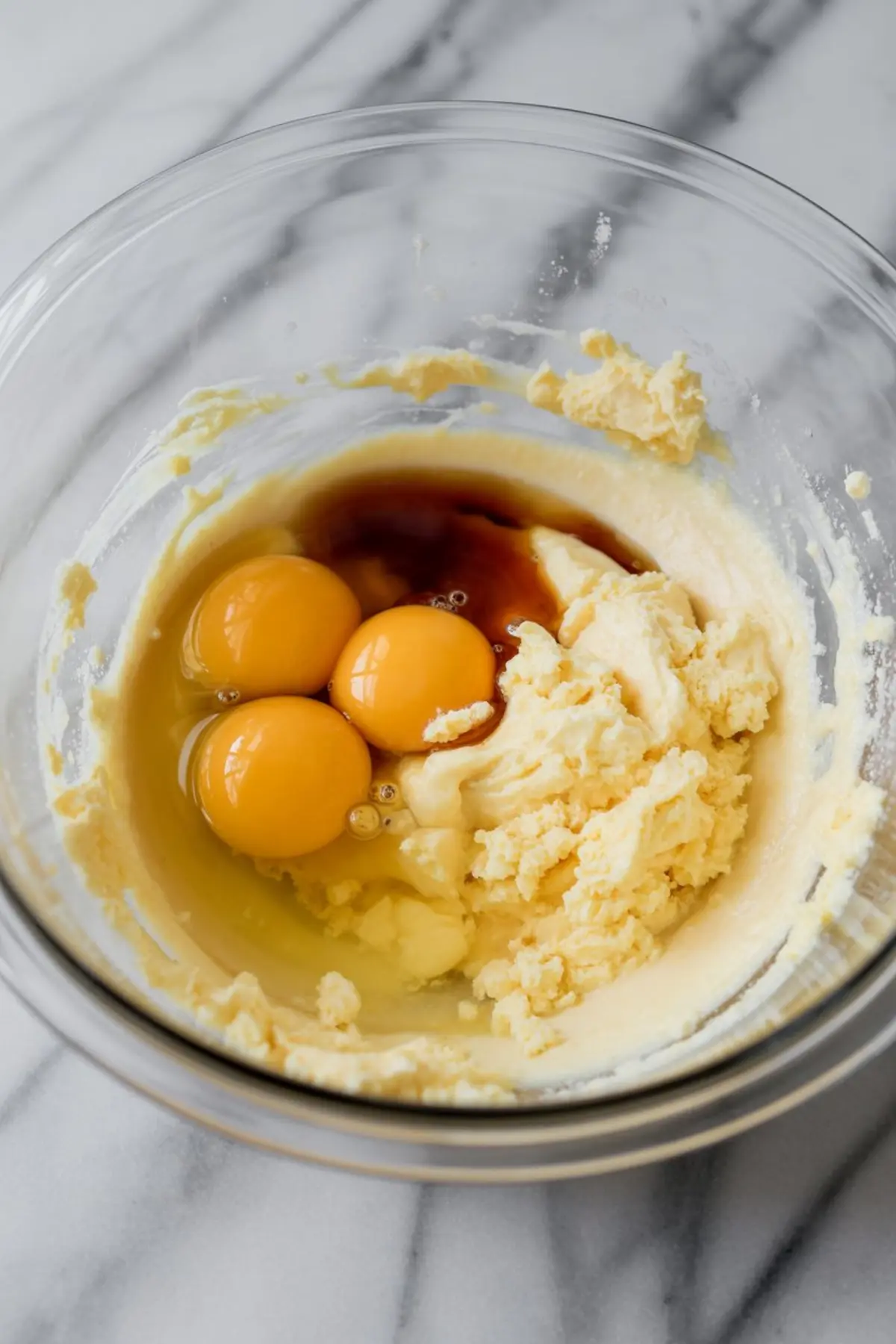Three raw eggs and vanilla extract rest atop partially creamed butter and sugar in a mixing bowl, ready to be blended into a cake batter.