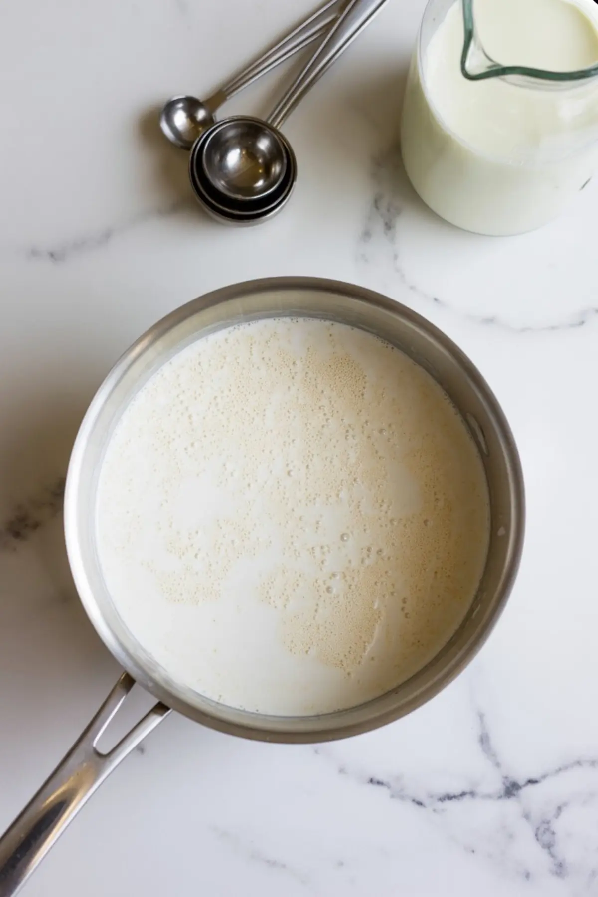 Stainless steel saucepan filled with simmering milk on a white marble countertop, with a glass pitcher of milk and measuring spoons nearby.
