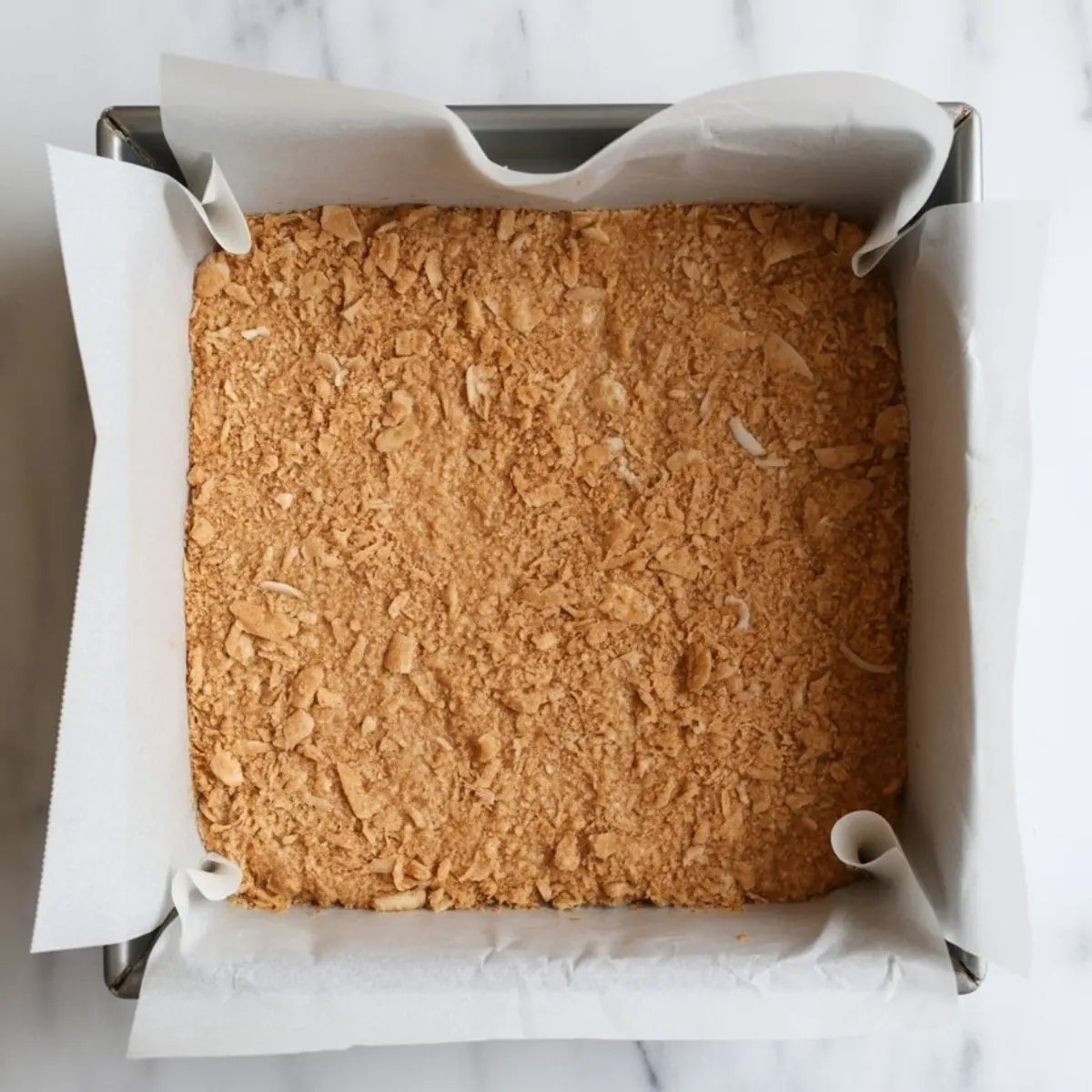 Square baking pan lined with parchment paper and filled with pressed graham cracker and coconut crust mixture ready for baking.
