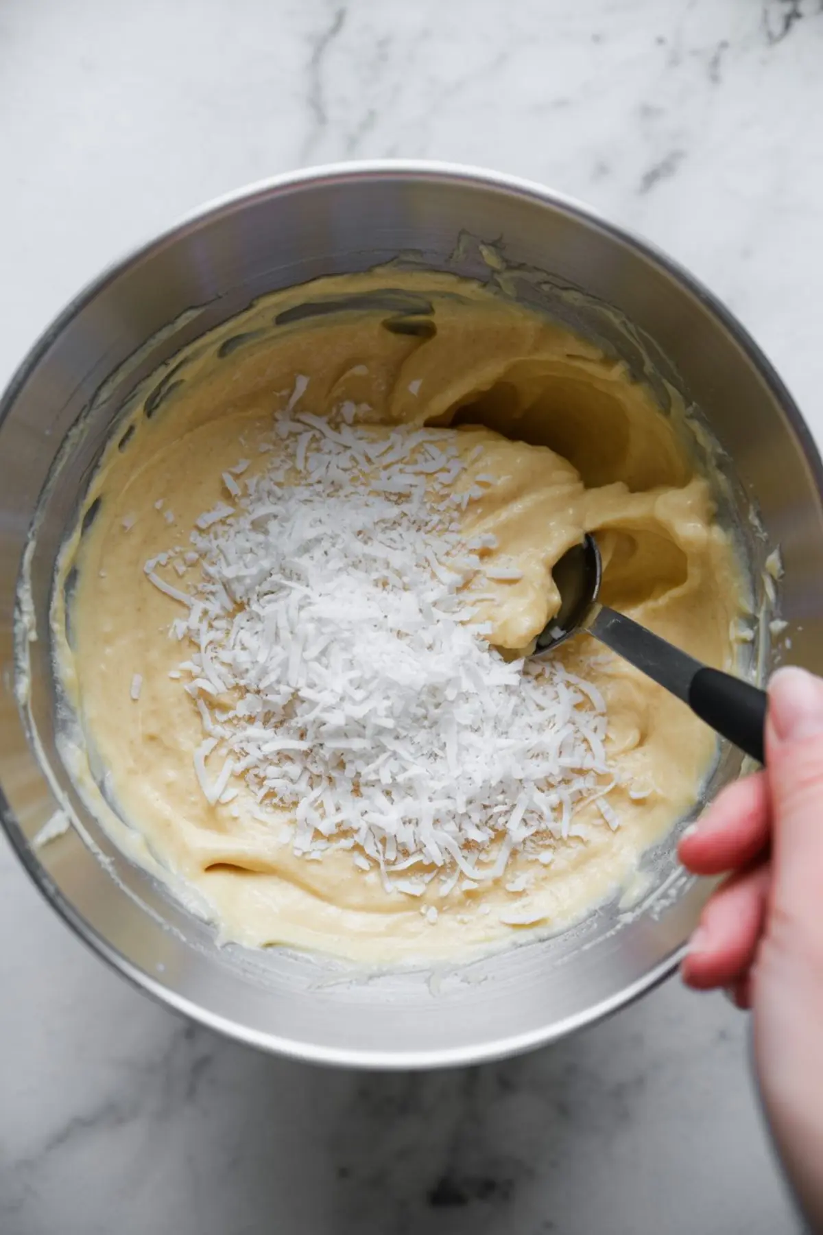 Shredded coconut added to a thick yellow cheesecake batter in a mixing bowl with a spoon.
