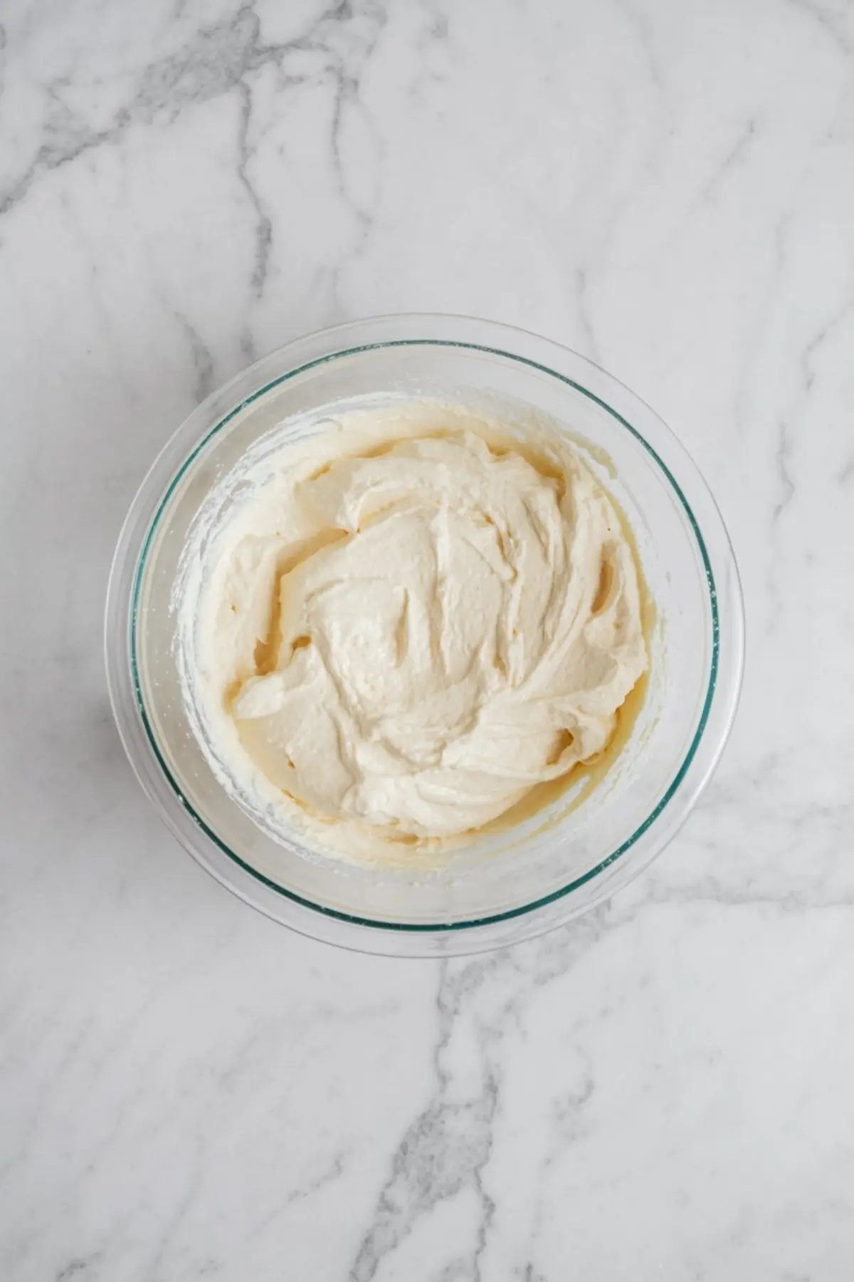 Creamy cheesecake batter mixed in a clear glass bowl on a white marble surface, showing smooth and thick texture.