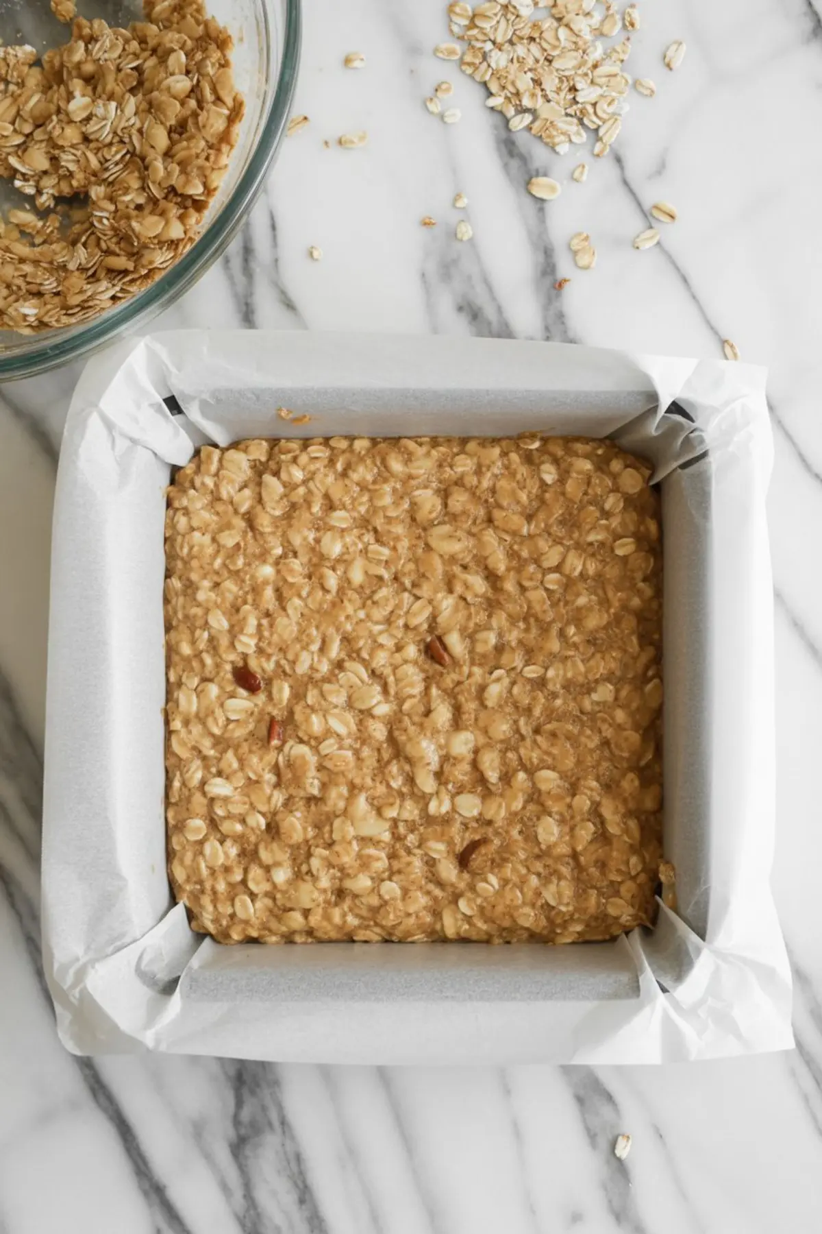 Unbaked oatmeal crust mixture pressed into a square baking pan lined with parchment paper, with extra oat mixture scattered on a marble countertop.