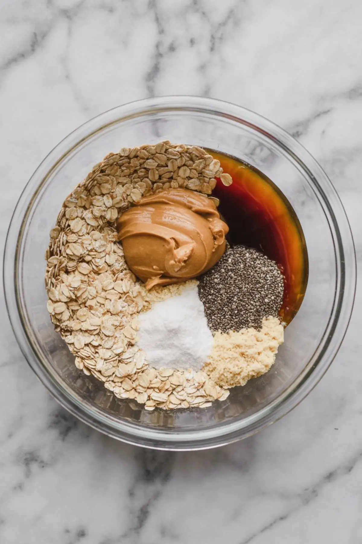 Top view of a glass bowl filled with dry and wet oatmeal cup ingredients including rolled oats, peanut butter, maple syrup, protein powder, chia seeds, baking powder, and salt, arranged before mixing.
