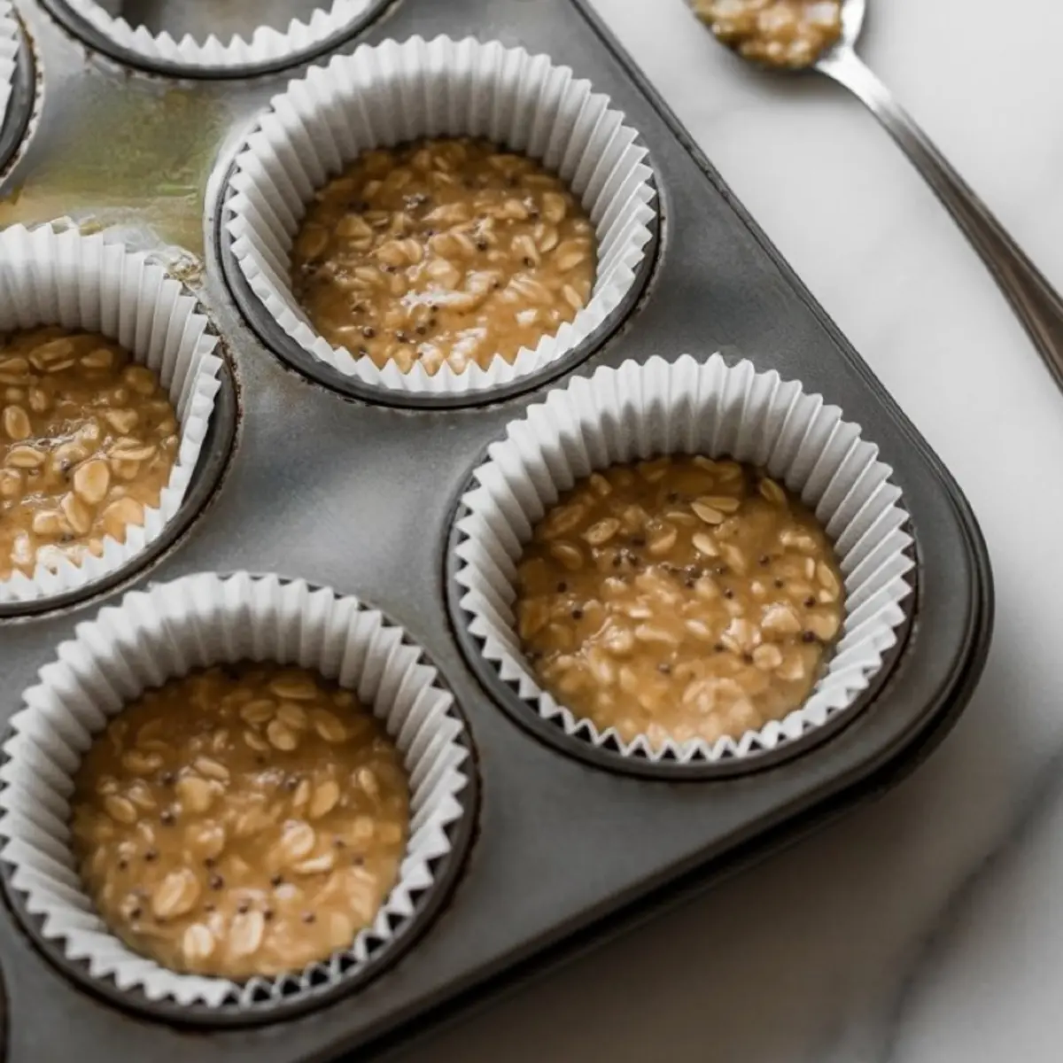 Close-up of muffin tin lined with white paper liners filled with uncooked oatmeal cup batter made from oats, peanut butter, and chia seeds, ready for baking or chilling.
