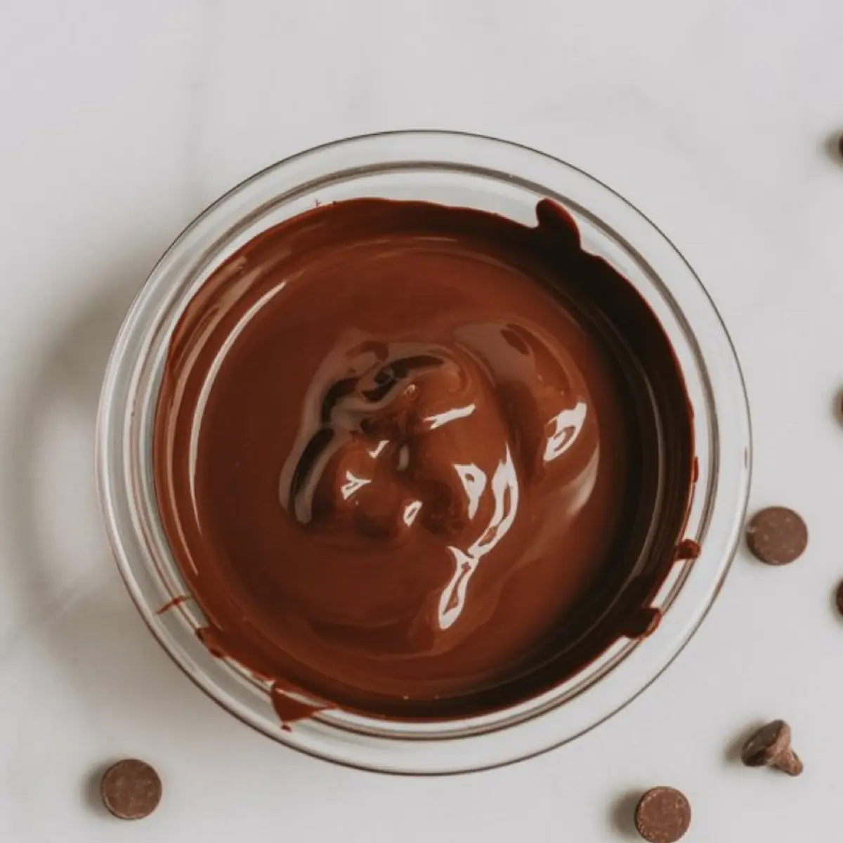 Glass bowl filled with smooth, melted dark chocolate ready for topping oatmeal cups, surrounded by scattered chocolate chips on a light marble surface.
