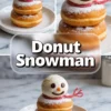 A festive donut snowman dessert made with two stacked powdered sugar donuts, topped with a vanilla ice cream scoop shaped like a snowman head. The snowman has chocolate chip eyes, a small orange candy nose, a curved chocolate mouth, and a red licorice scarf. Styled on a white plate over a marble background, the image includes close-ups and full views with a bold “Donut Snowman” text overlay.