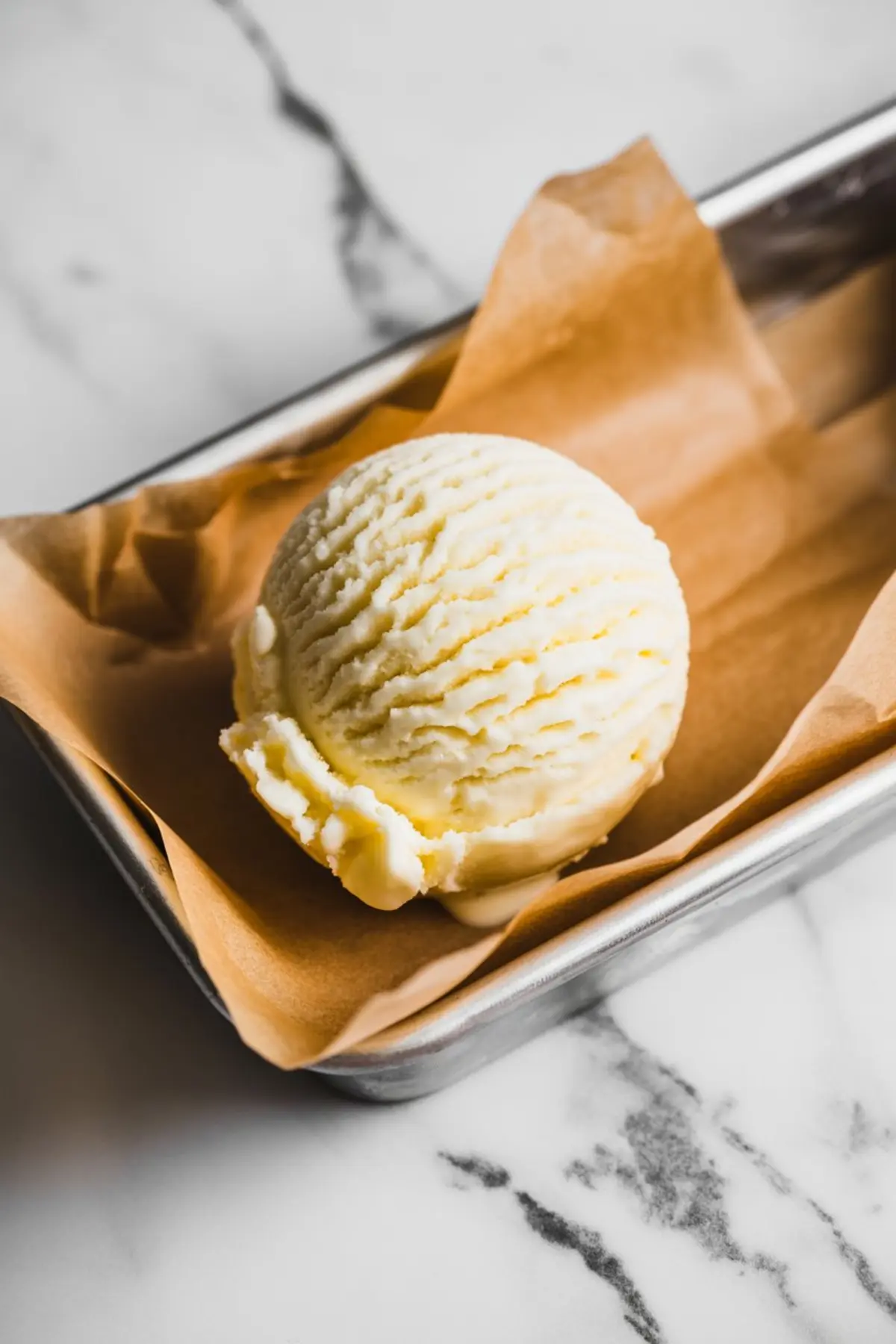 A single scoop of vanilla ice cream placed in a small metal tray lined with brown parchment paper, set on a marble surface. The ice cream has a creamy, smooth texture with visible scoop lines and slightly melting edges.
