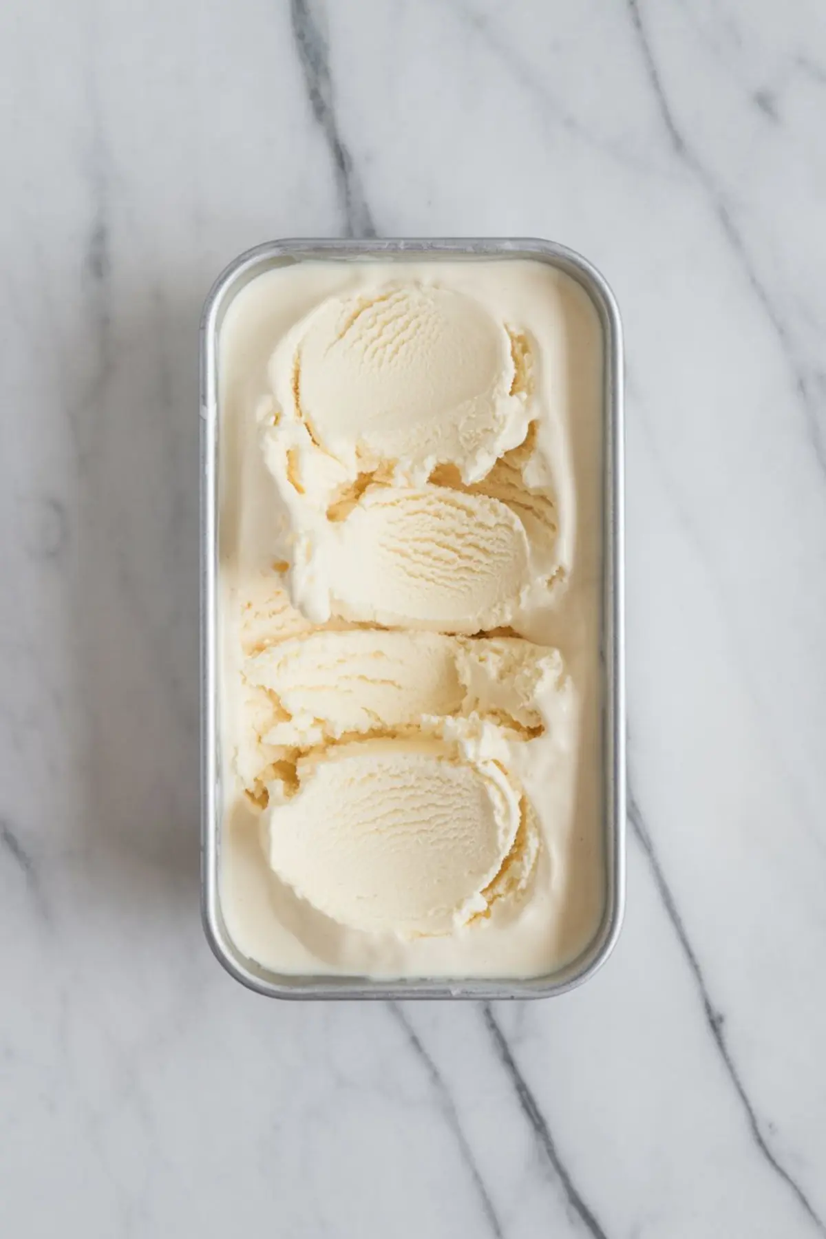 A metal container filled with vanilla ice cream, showing multiple smooth scoops with rich, creamy texture. The container is placed on a light marble surface, emphasizing the classic dessert base.
