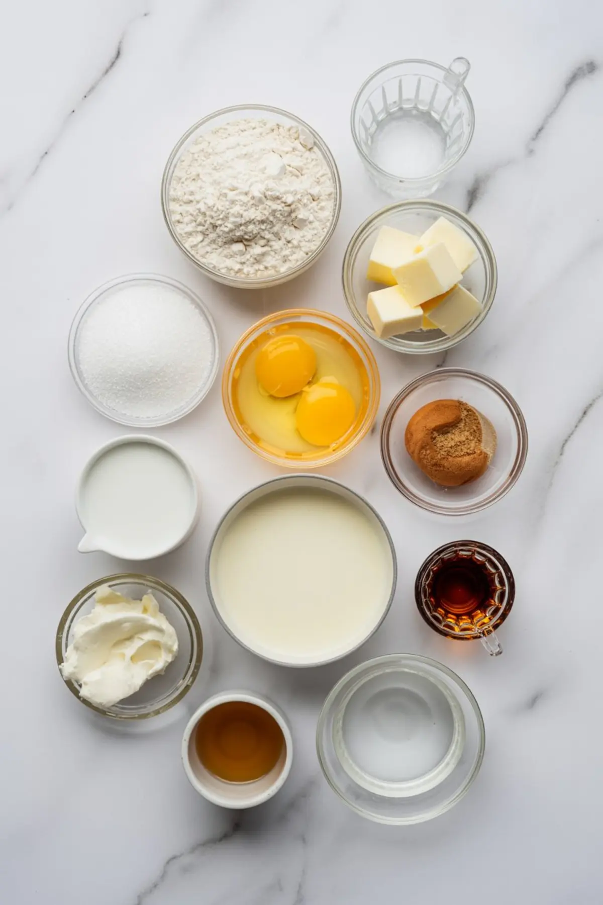 Baking ingredients arranged in small bowls on a white marble background, including eggs, butter, flour, sugar, milk, vanilla, cream, and spices.
