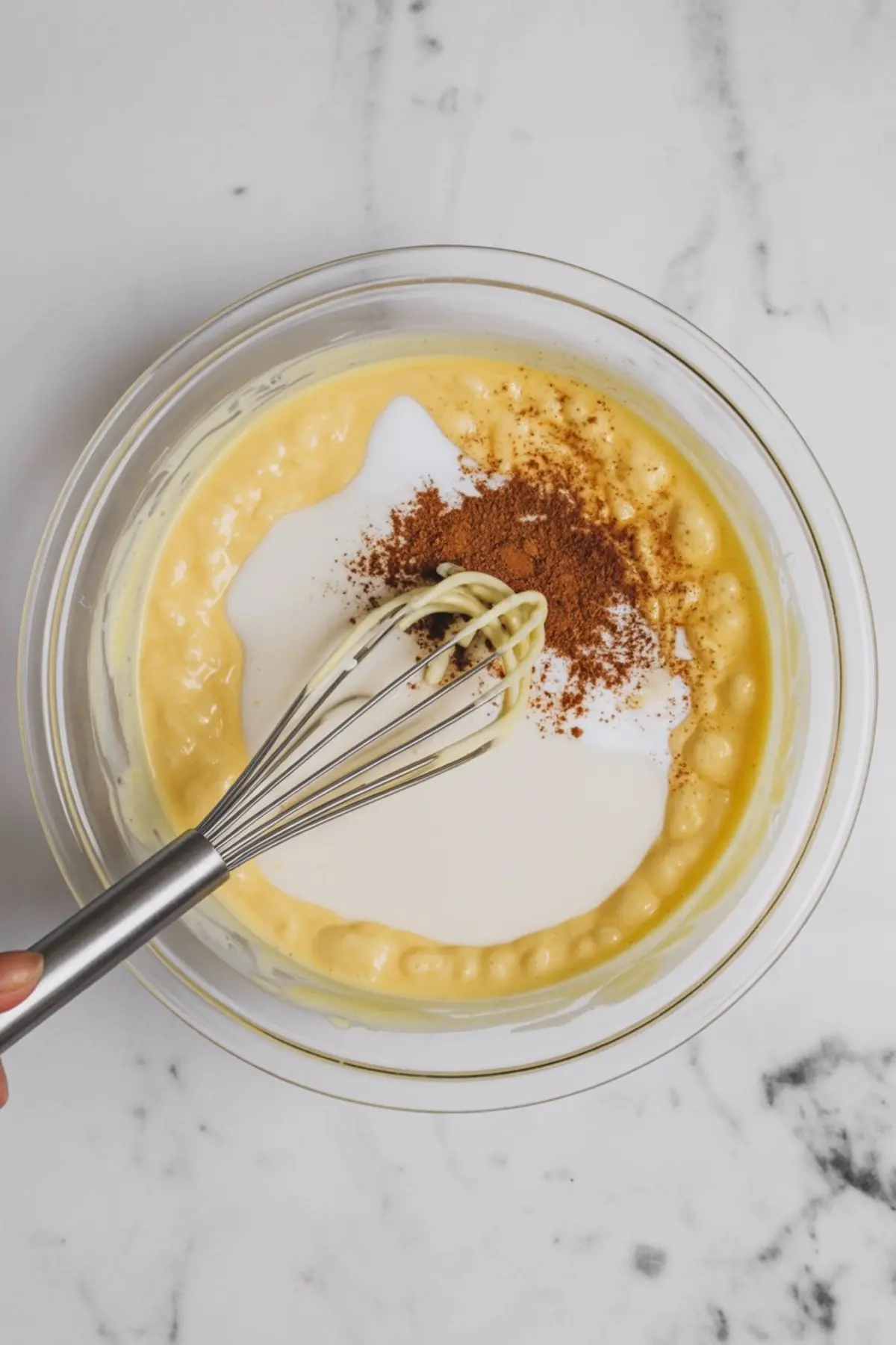 Close-up of a glass bowl filled with thick eggnog pudding base, topped with cream and ground cinnamon, with a metal whisk partially submerged in the mixture, placed on a marble background.
