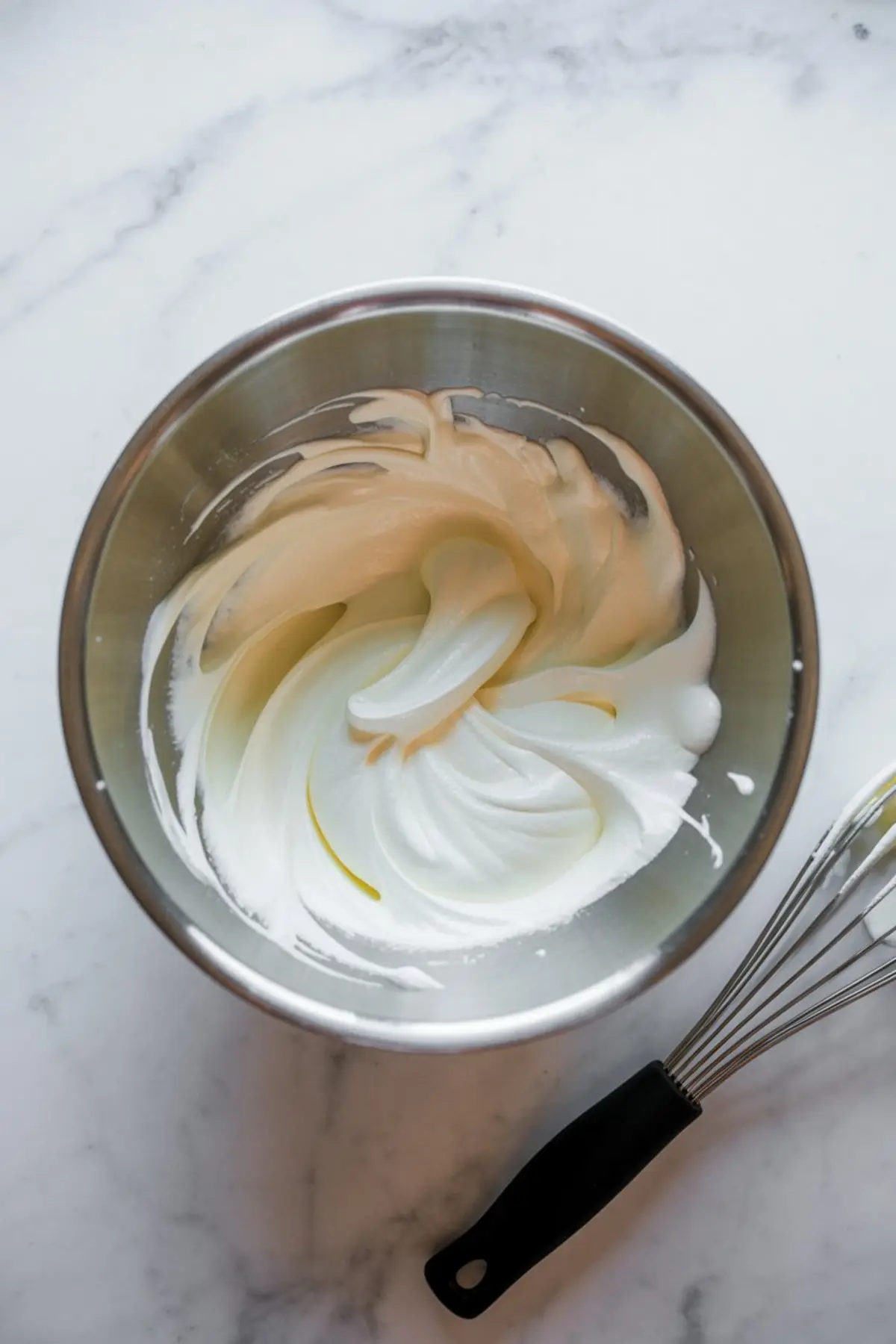 Whipped egg whites in a metal mixing bowl with a soft peak consistency, next to a wire whisk on a marble surface.
