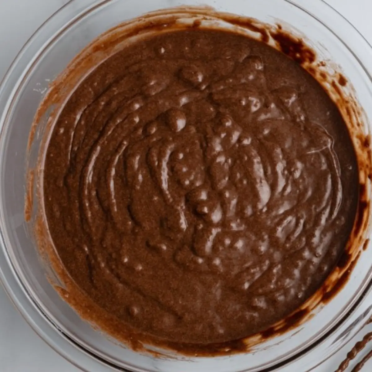 Glossy chocolate brownie batter in a clear glass mixing bowl, showing a smooth texture with visible air pockets.