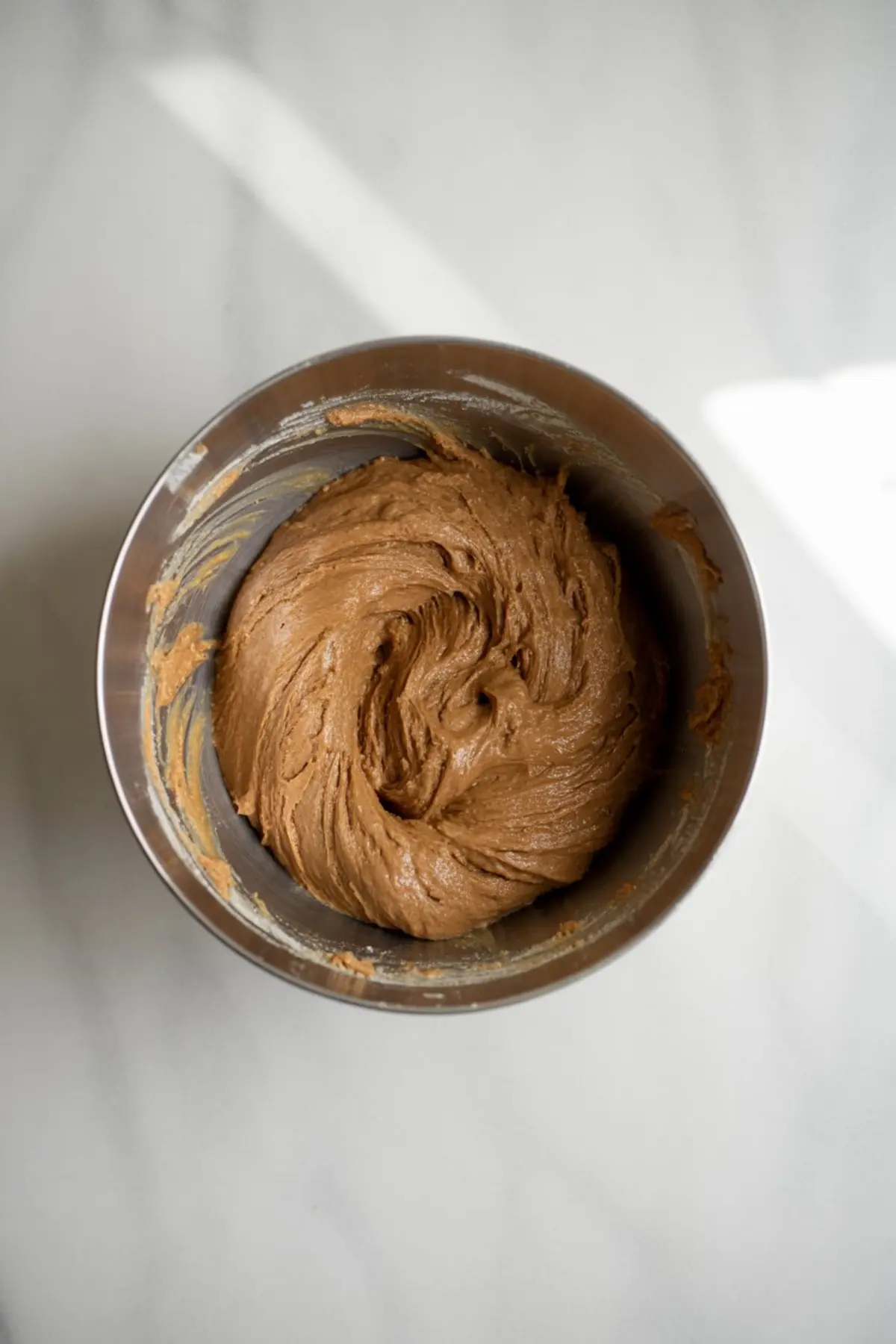 Creamy gingerbread cookie dough swirled in a stainless steel mixing bowl on a white marble countertop.