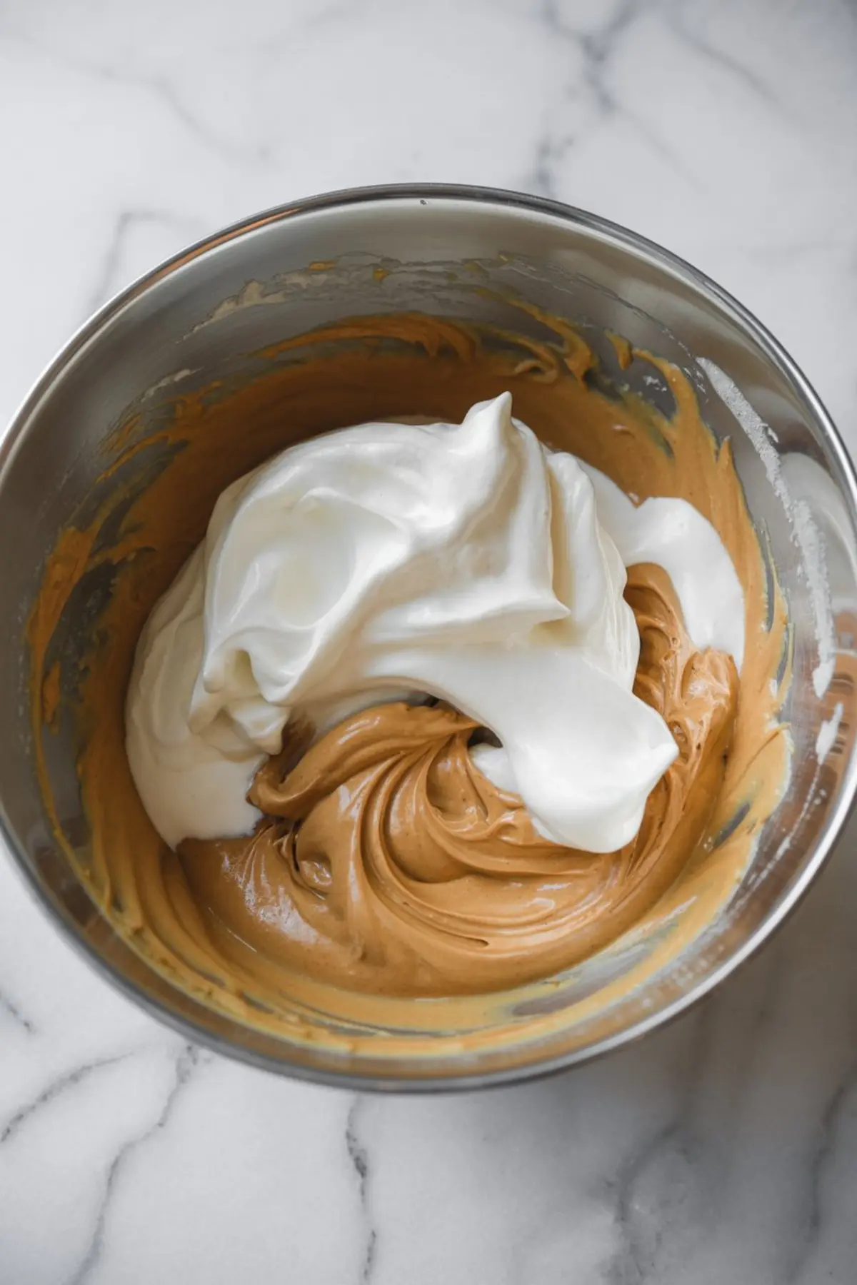 Partially folded meringue and spiced mousse base in a bowl, showing a fluffy white topping on top of brown gingerbread mixture.