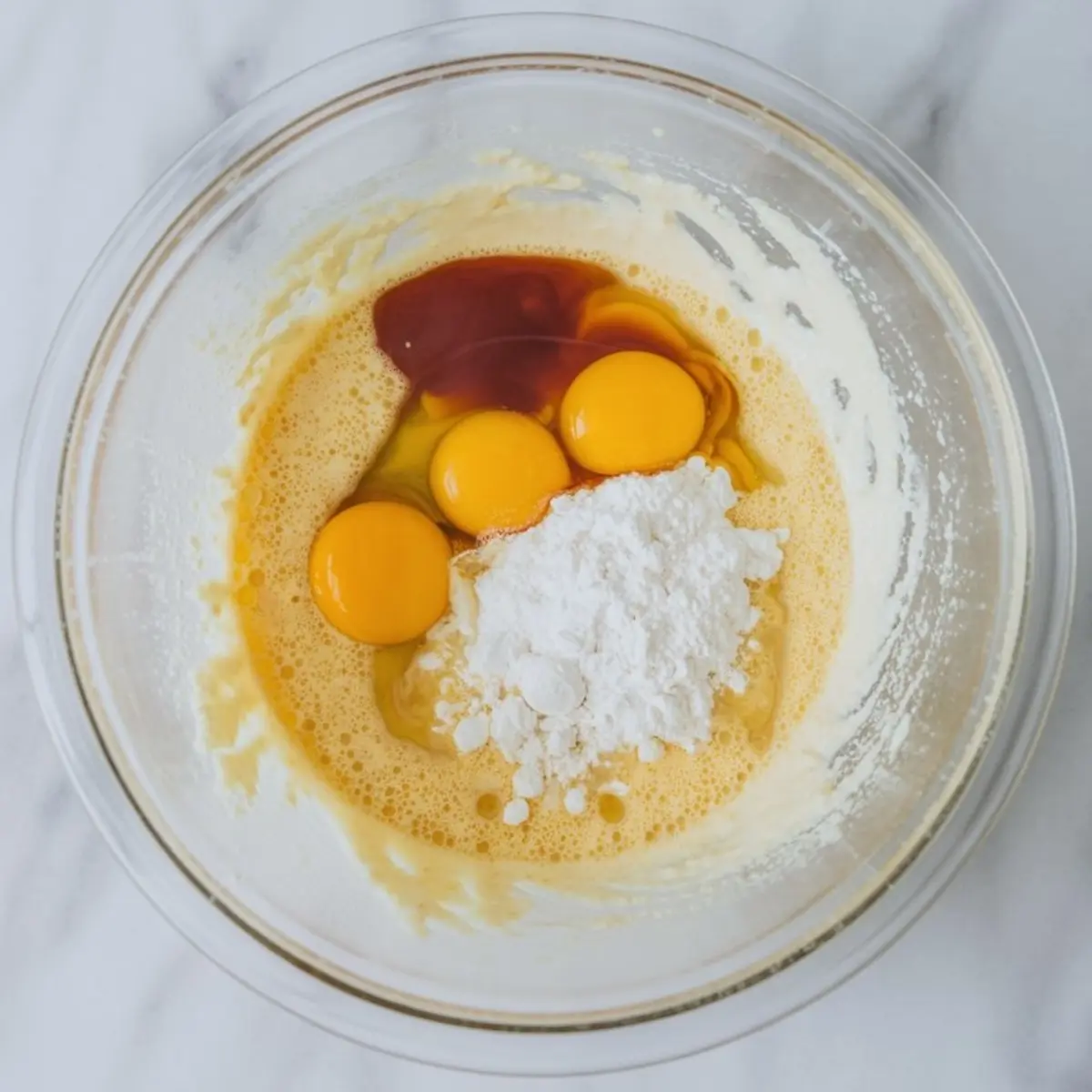 Bowl of partially mixed ingredients including three egg yolks, powdered sugar, and vanilla extract sitting on a creamy whipped base, ready to be blended for baking.
