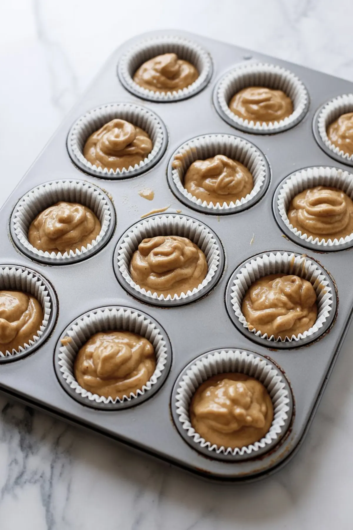Twelve cupcake liners filled with thick gingerbread batter in a metal muffin tray, set on a white marble background, ready to bake.
