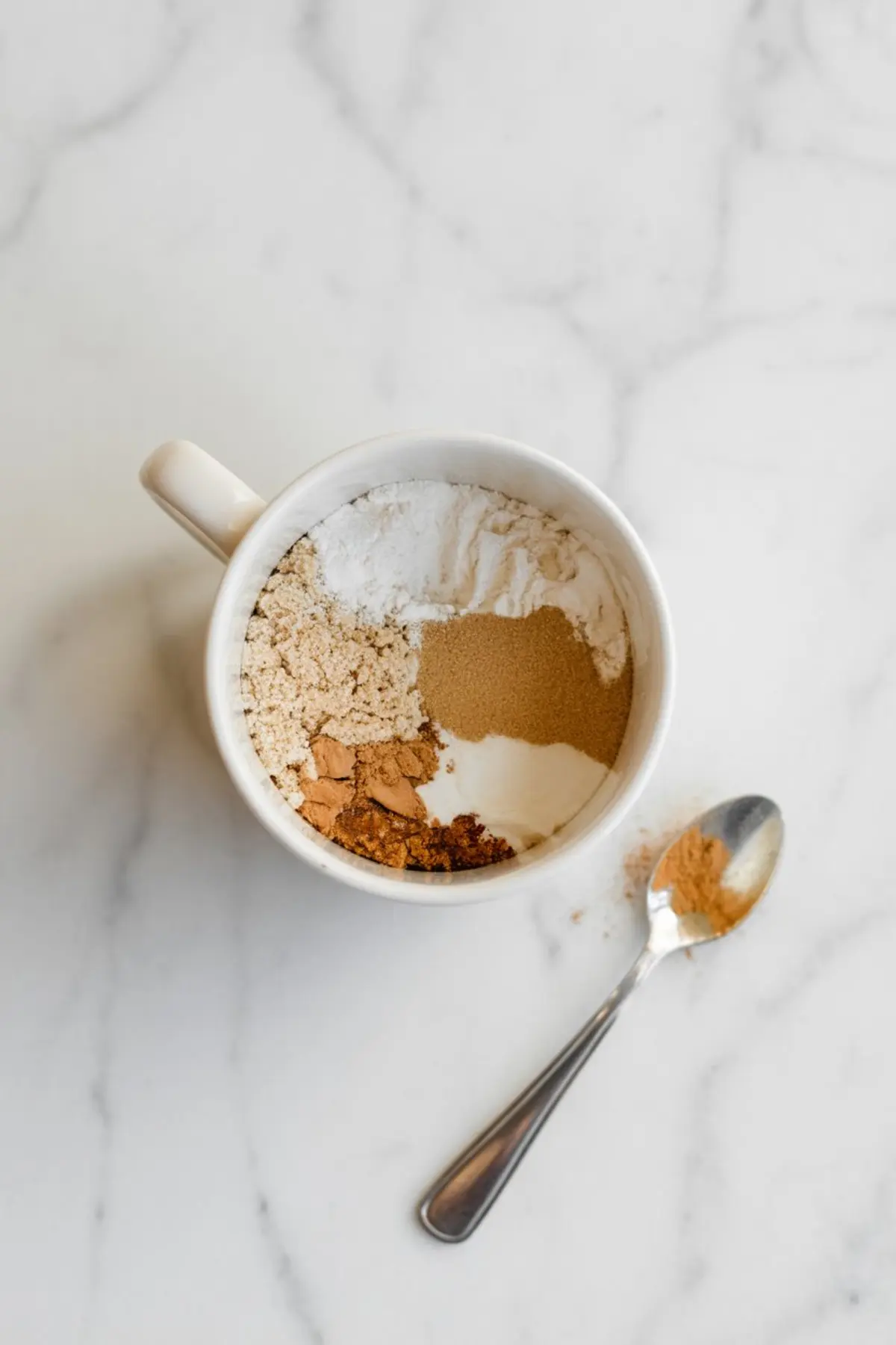 Dry ingredients for gingerbread mug cake including flour, brown sugar, baking powder, and spices arranged separately in a white mug with a spoon holding cinnamon beside it.
