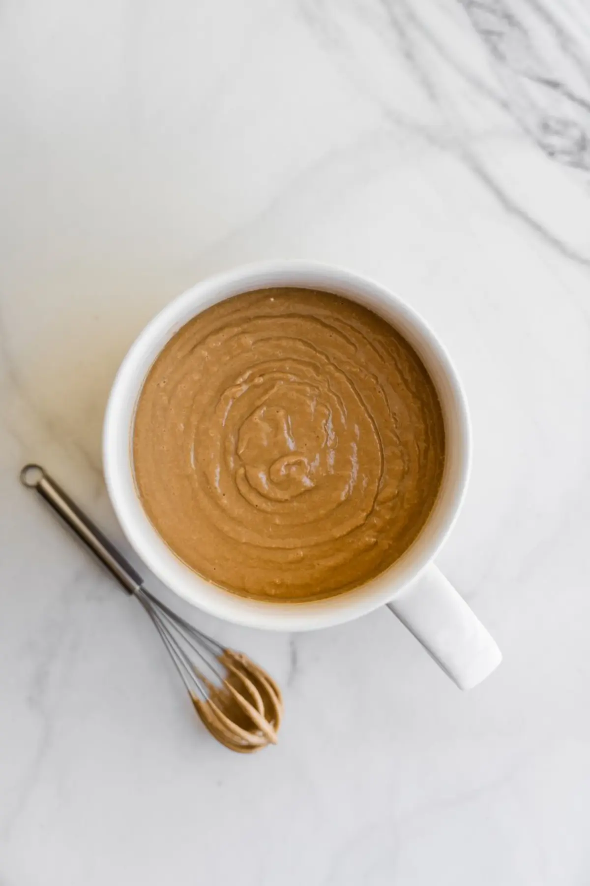Mixed gingerbread batter in a white mug with a spiral texture on top, sitting on a marble surface next to a small metal whisk coated in batter.
