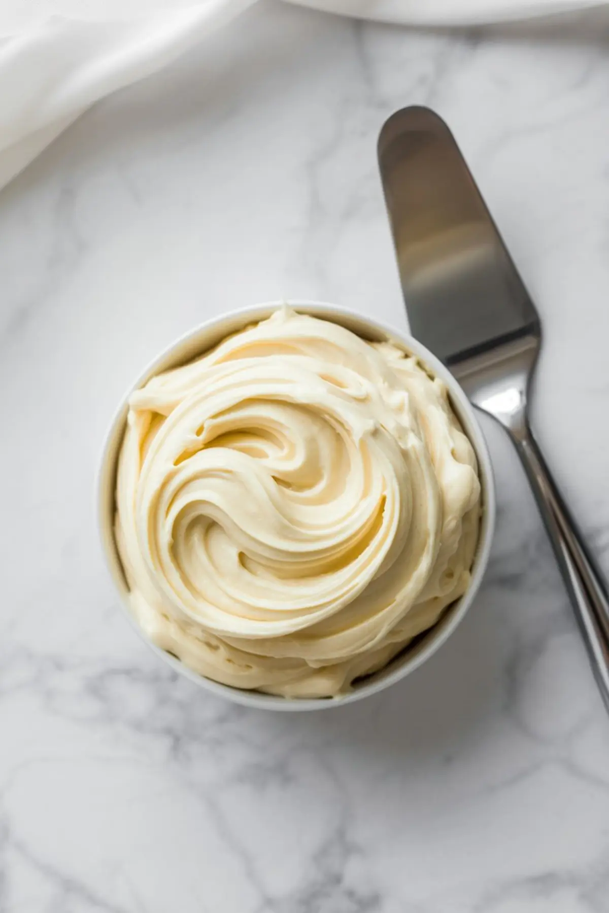 Whipped cream cheese frosting with smooth swirls in a small white bowl, placed beside a metal offset spatula on a marble countertop.
