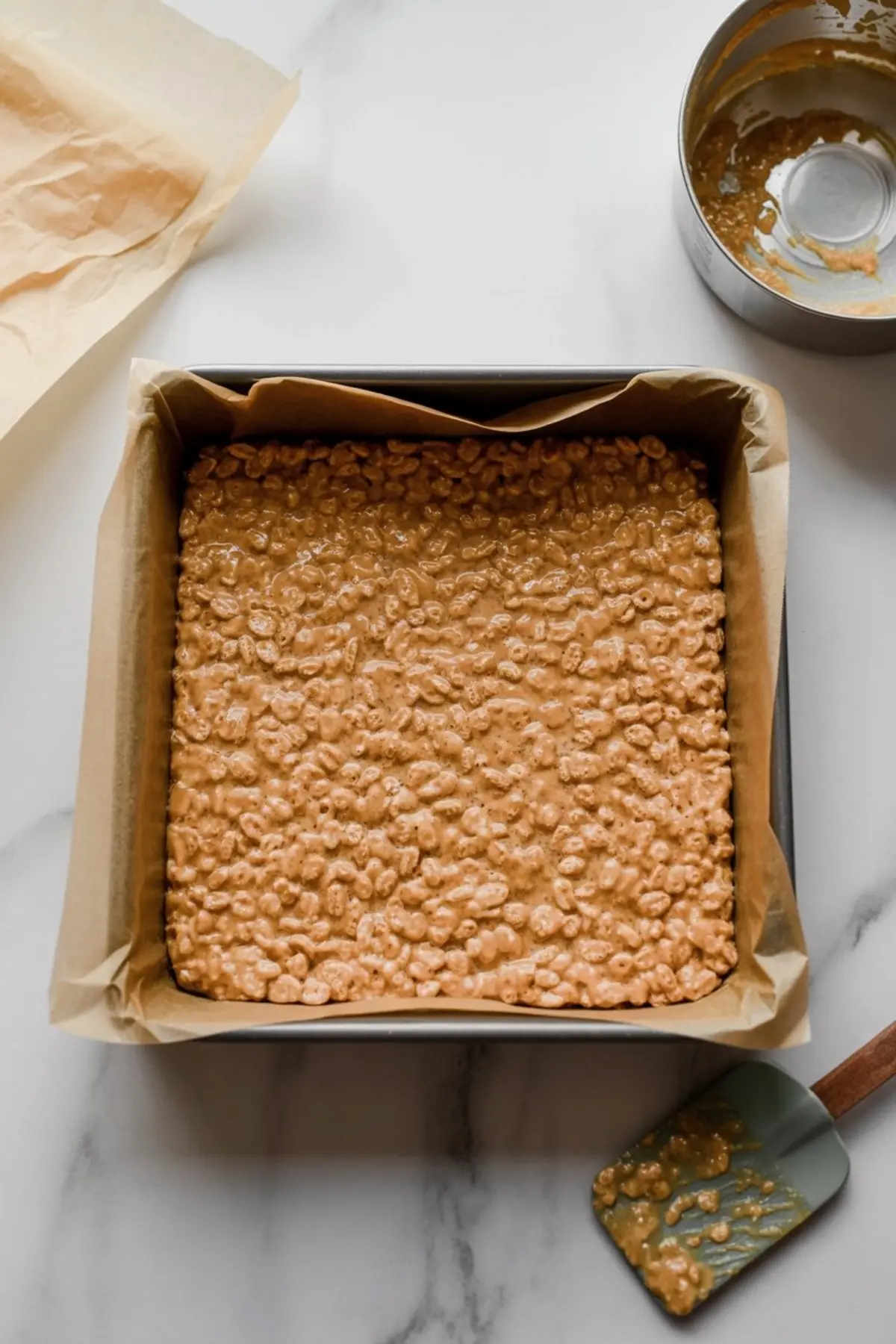 A baking pan lined with parchment paper holds a pressed mixture of puffed rice cereal coated in a glossy gingerbread syrup blend, ready for slicing.