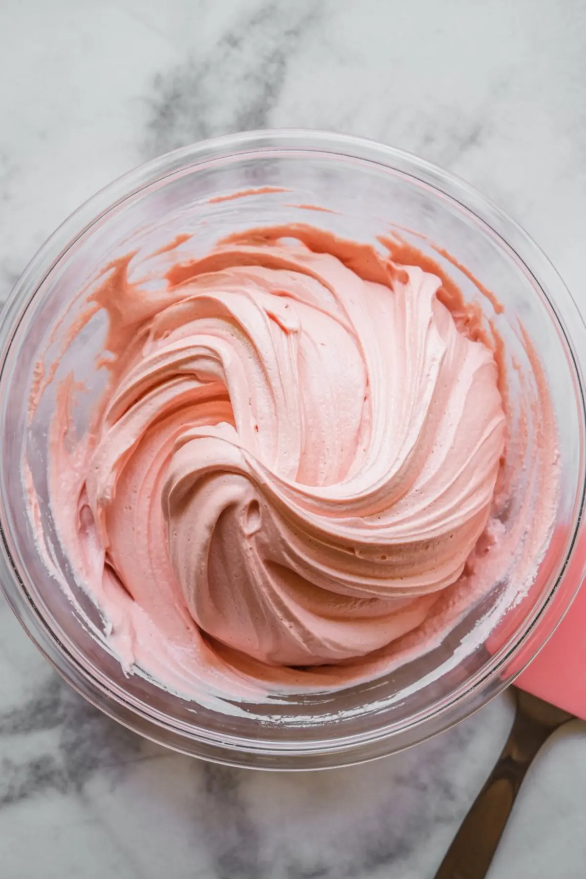 Image shows a bowl of pink whipped cheesecake mixture with smooth, creamy swirls in a clear glass bowl on a marble surface.
