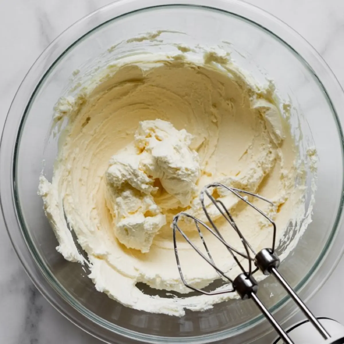Image shows a bowl of whipped cream cheese mixture being blended with electric mixer beaters resting inside the glass bowl on a marble countertop.
