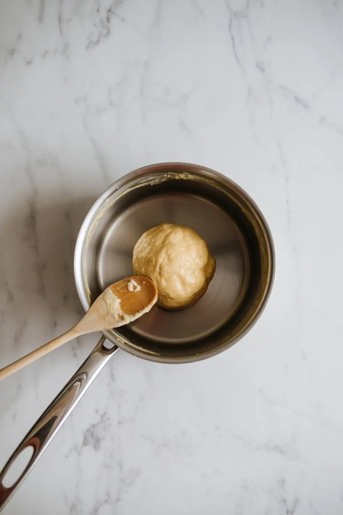 Cooked choux pastry dough formed into a ball in a stainless steel saucepan with a wooden spoon, showing the cooked pâte à choux base used for making cream puffs and éclairs.