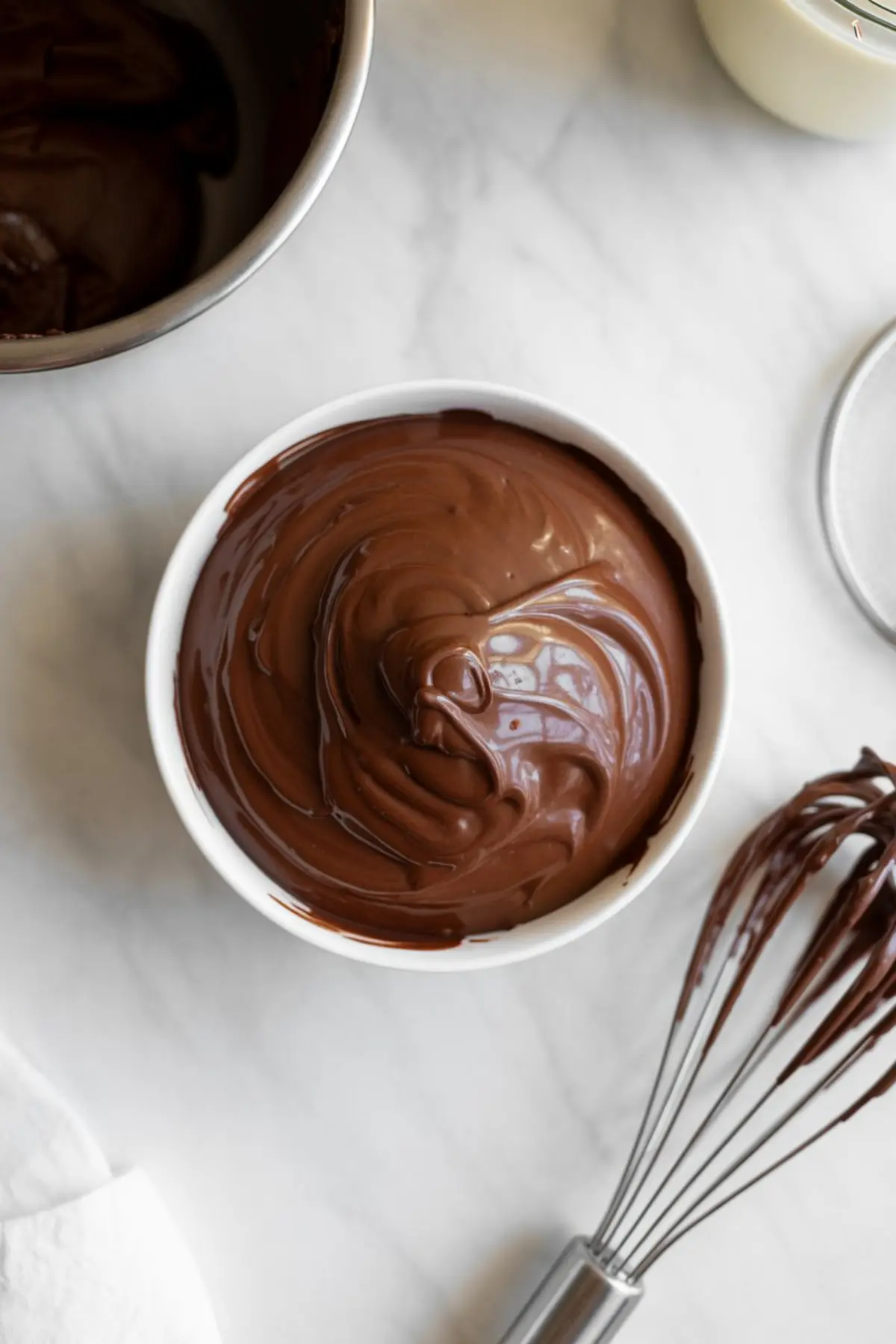 Bowl filled with smooth, swirled chocolate pastry cream next to a chocolate-covered whisk, set on a light marble countertop for a rich dessert filling preparation.