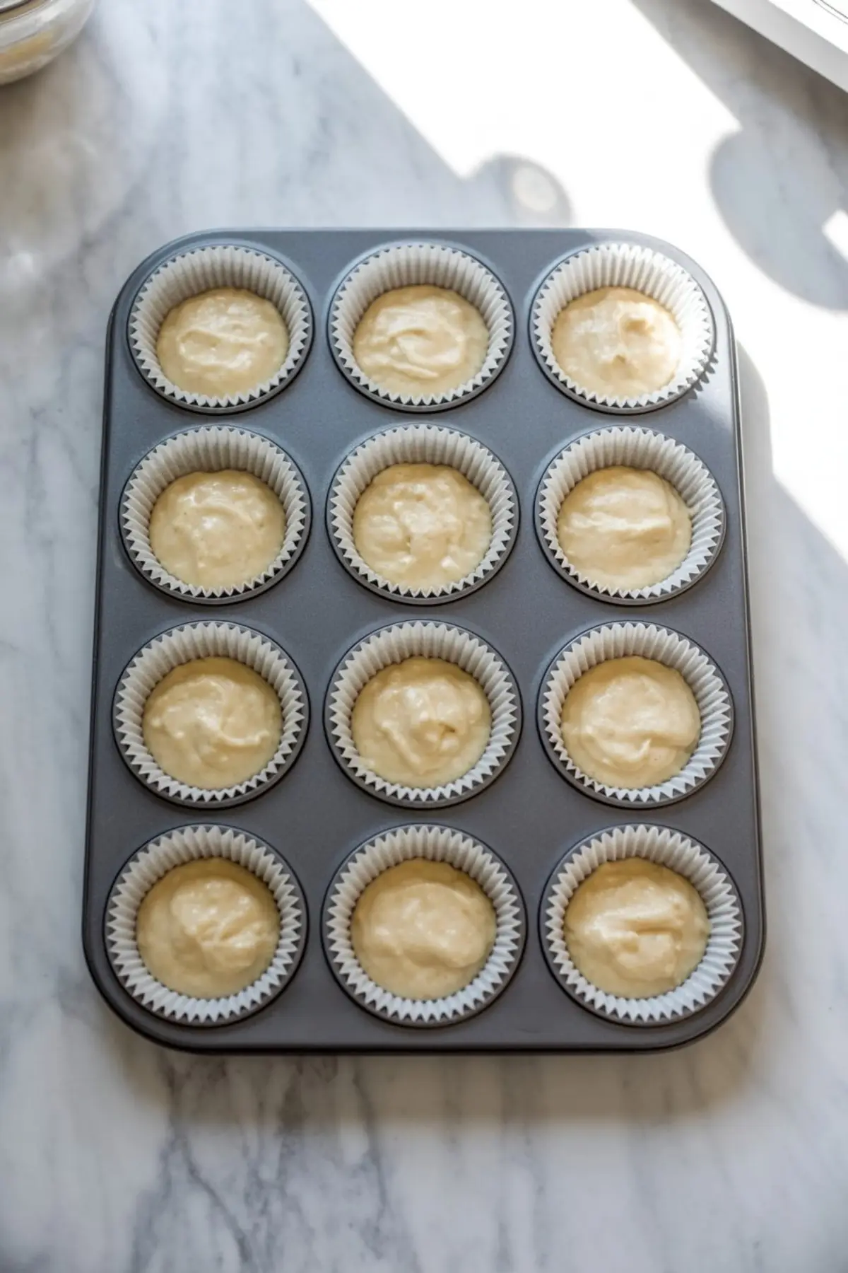 Cupcake batter portioned into paper liners in a 12-cup muffin tin, ready for baking, positioned under natural light on a marble kitchen counter.