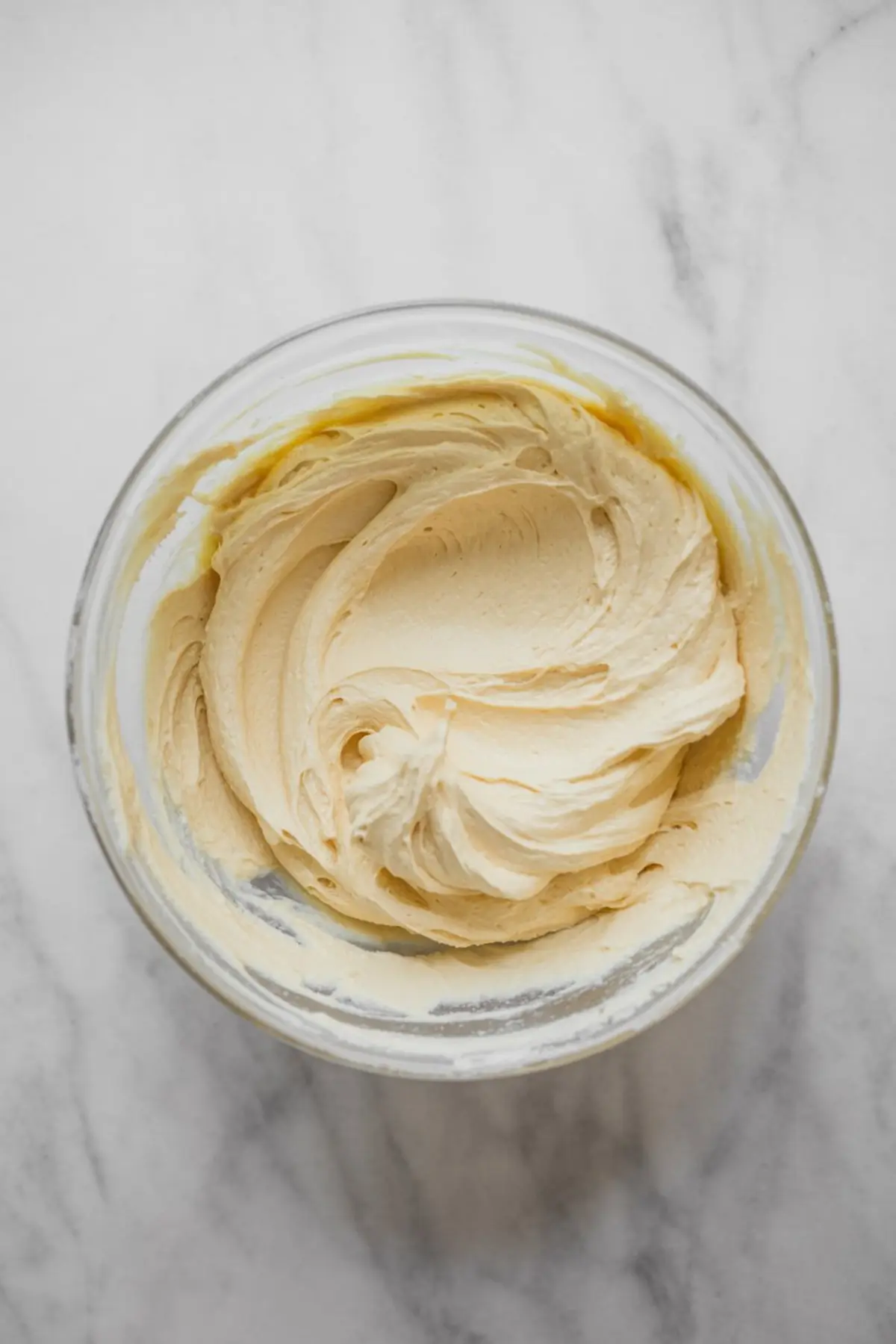 Creamy vanilla buttercream frosting whipped to a smooth texture in a glass bowl, prepared for decorating cupcakes on a white marble background.