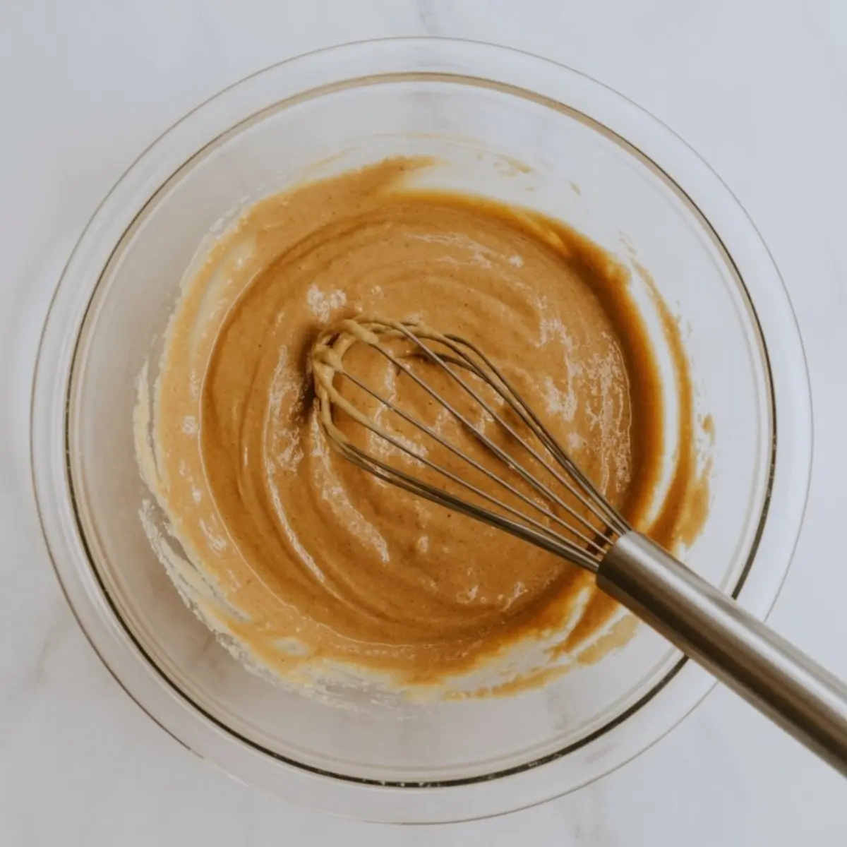 A glass bowl shows a blended maple and brown butter mixture with a whisk resting in the center.
