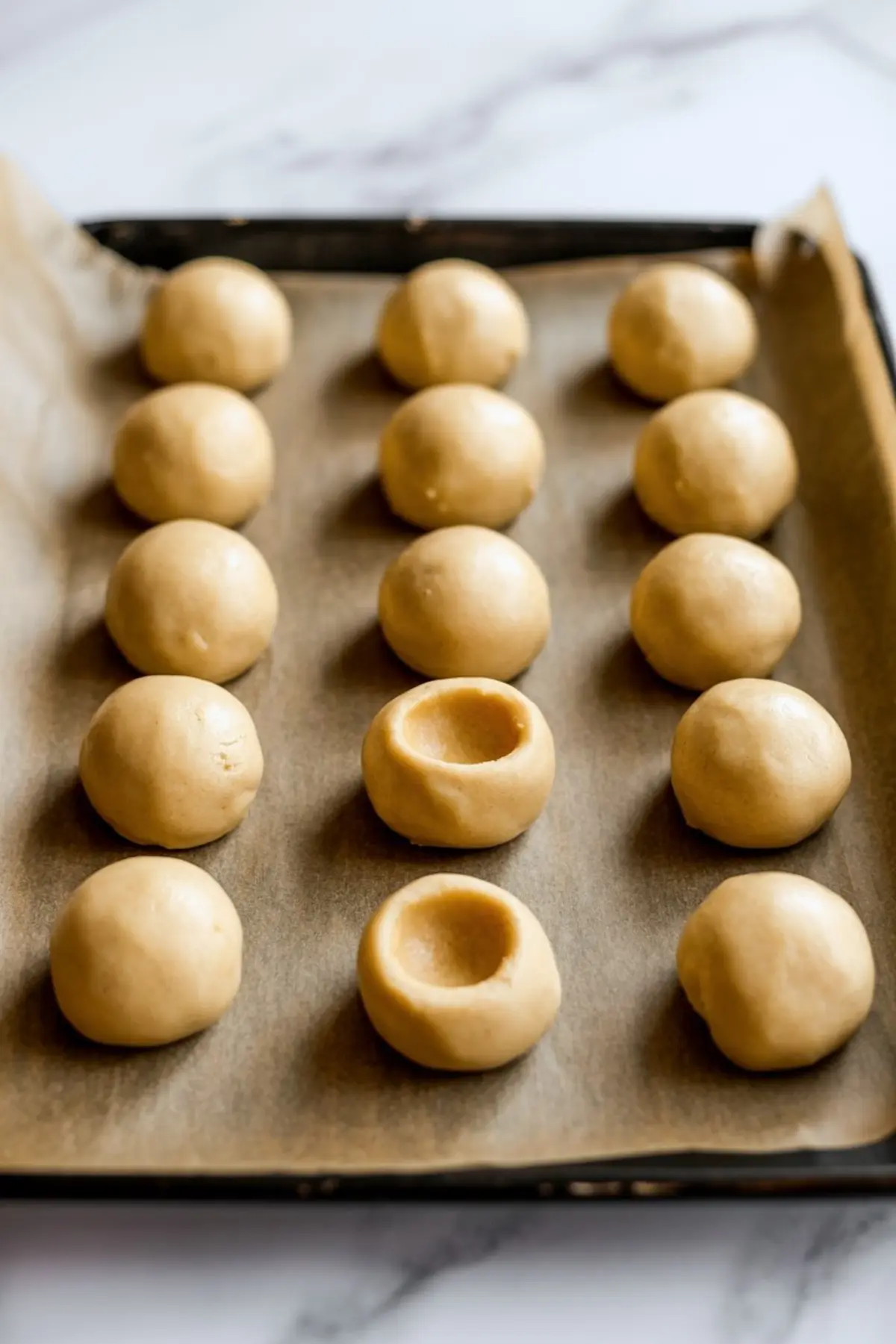 Raw cookie dough balls arranged in neat rows on a parchment-lined baking tray, some pressed in the center for thumbprint cookies.
