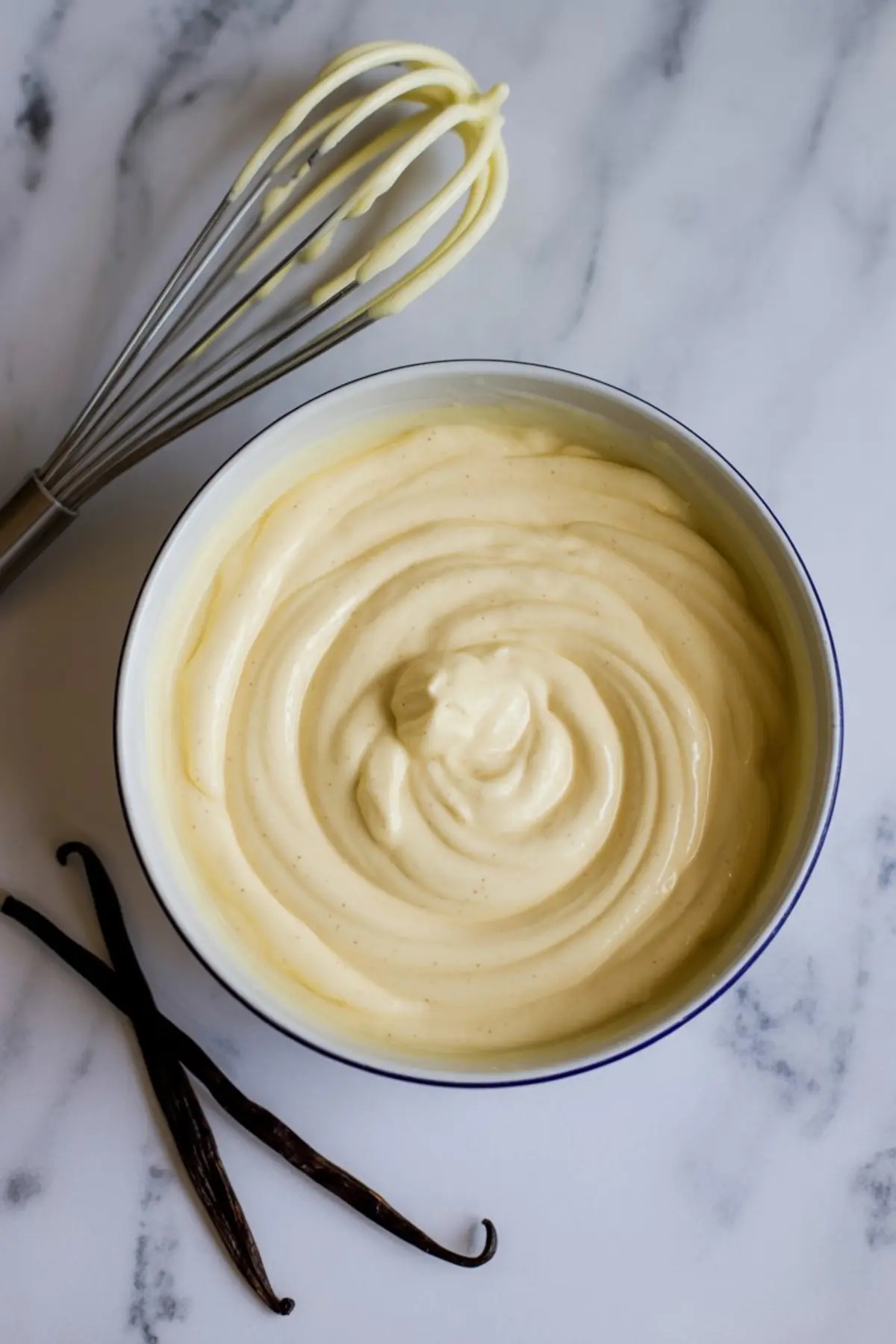 A mixing bowl of whipped vanilla pastry cream with visible flecks of vanilla bean, styled beside whole vanilla pods and a whisk on a marble counter.
