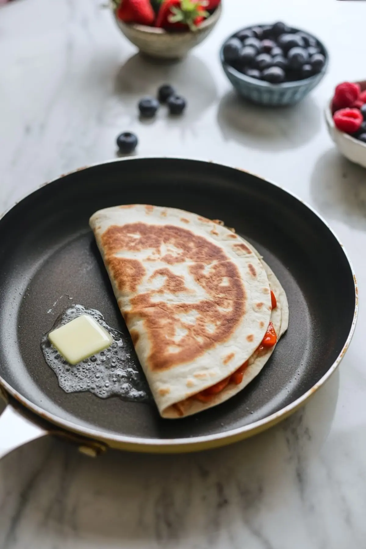 A folded Nutella-stuffed quesadilla with a golden-brown crust sizzles in a skillet beside a melting square of butter, with bowls of fresh berries in the background.