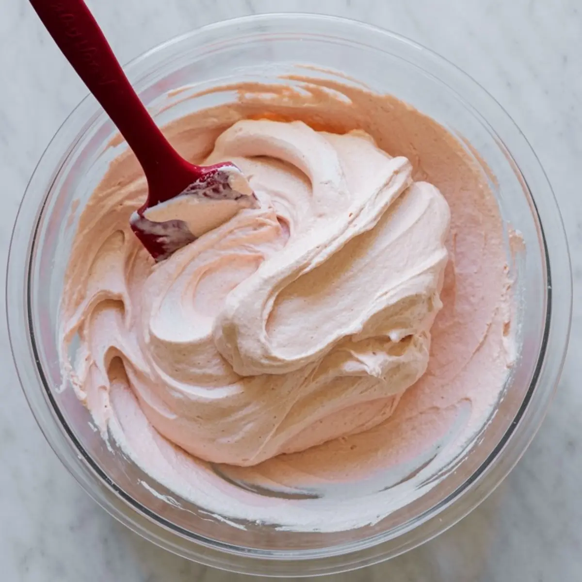 Glass bowl filled with smooth pink whipped peppermint cheesecake mousse being mixed with a red spatula on a marble surface.
