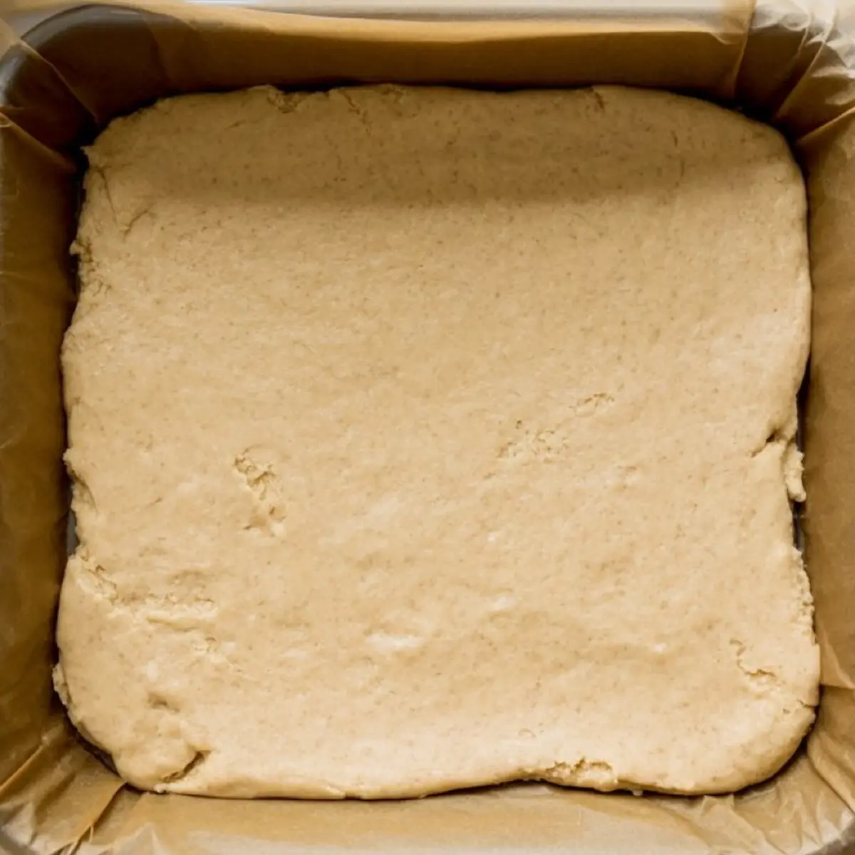 Baking pan lined with parchment paper filled with smooth cookie dough evenly pressed into the bottom.
