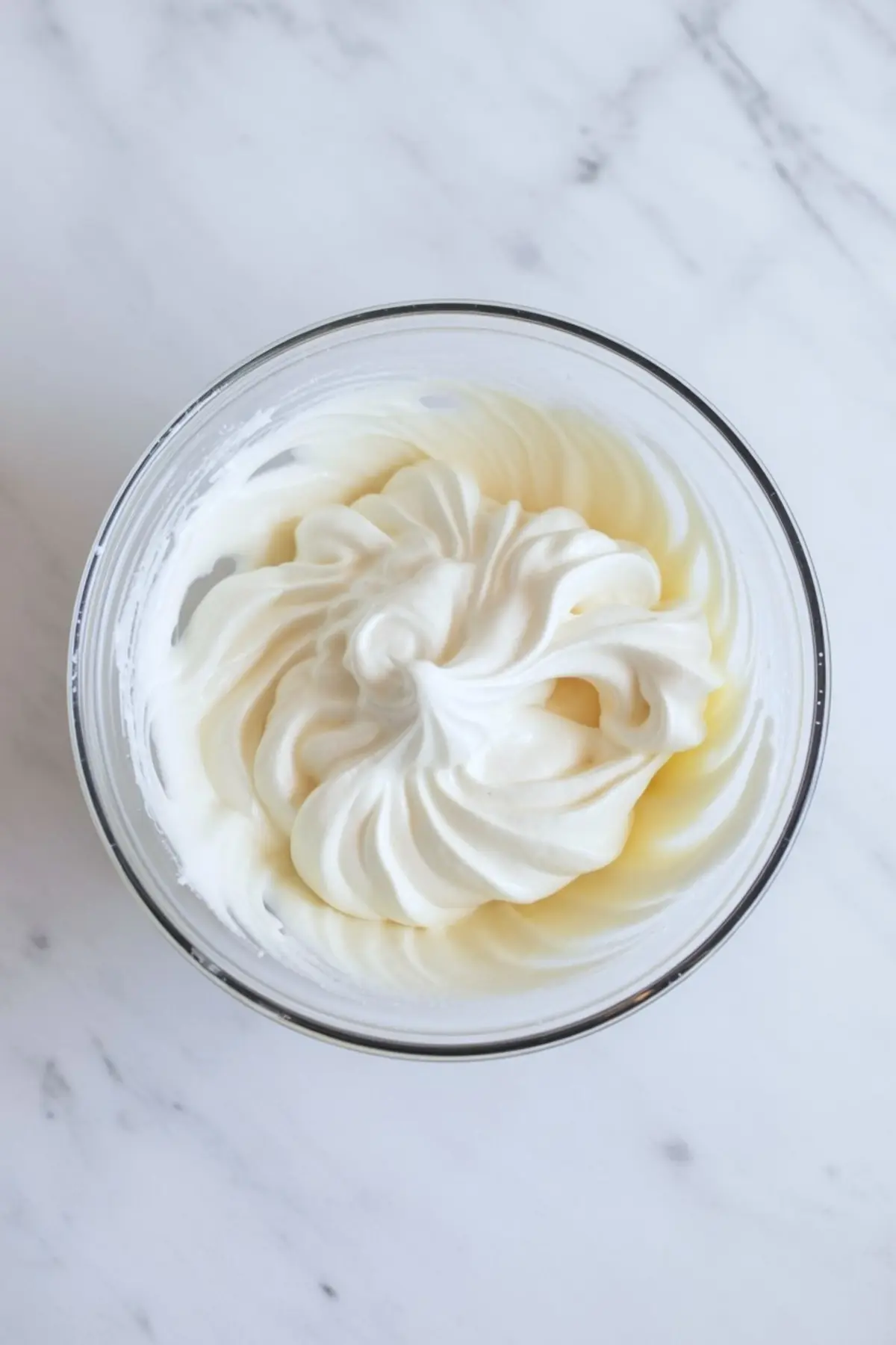 Freshly whipped cream with soft peaks in a glass bowl, sitting on a light marble countertop.
