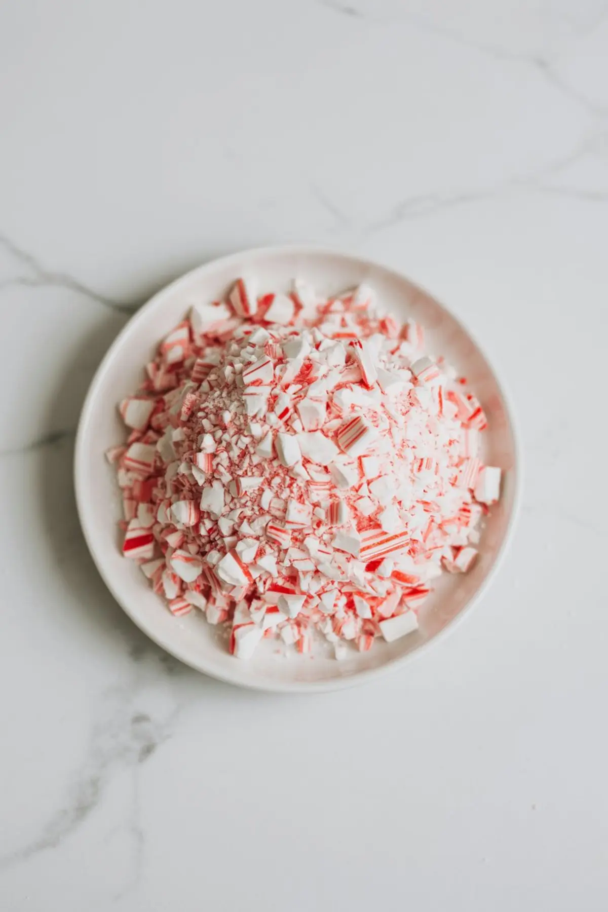 Crushed peppermint candy pieces in red and white colors piled on a white plate over a marble surface, ideal for holiday dessert toppings and festive drink garnishes.
