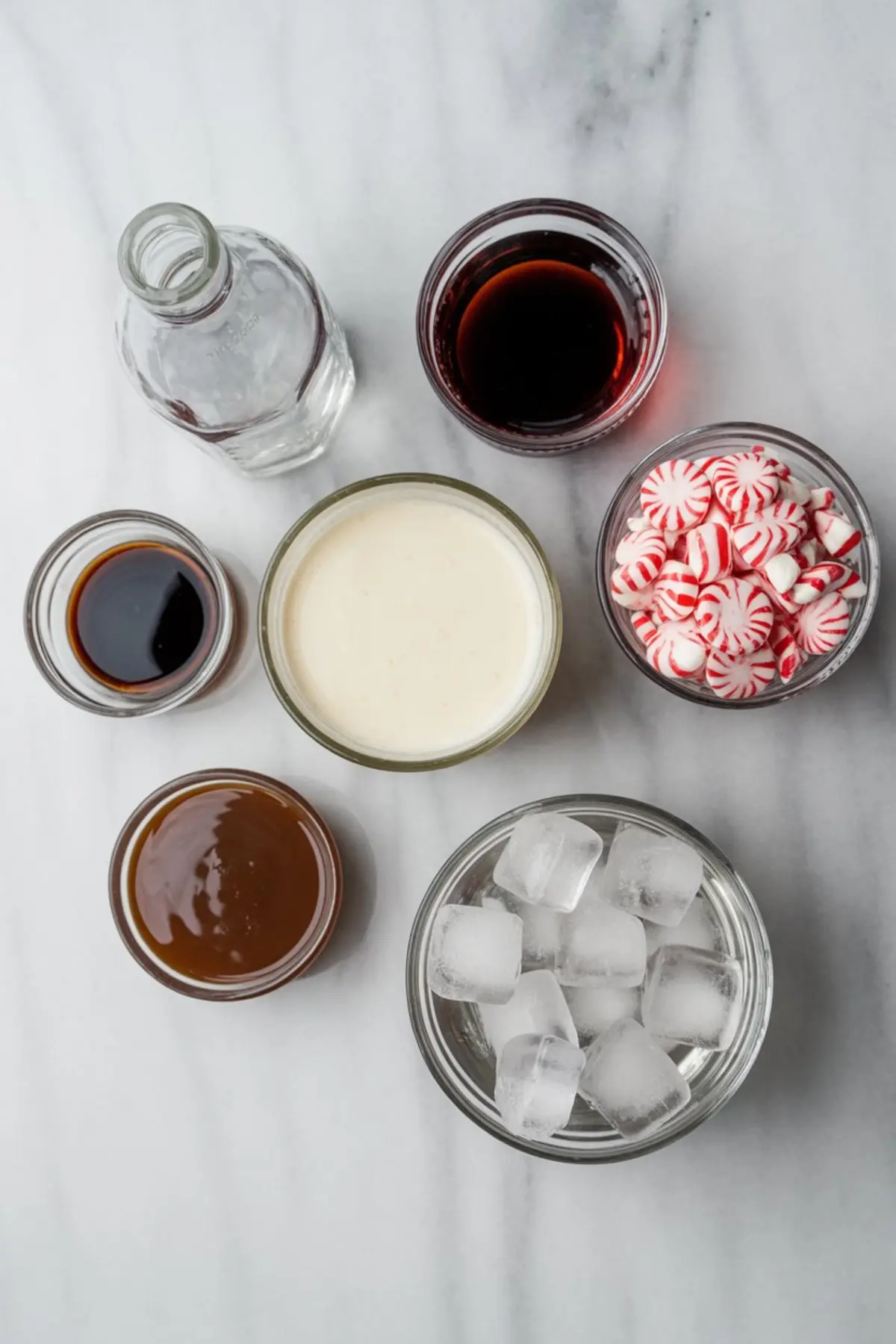 Flat lay of ingredients on a marble background including peppermint candies, ice cubes, cream, coffee liqueur, vodka, vanilla extract, and caramel sauce, arranged for making a peppermint cocktail.
